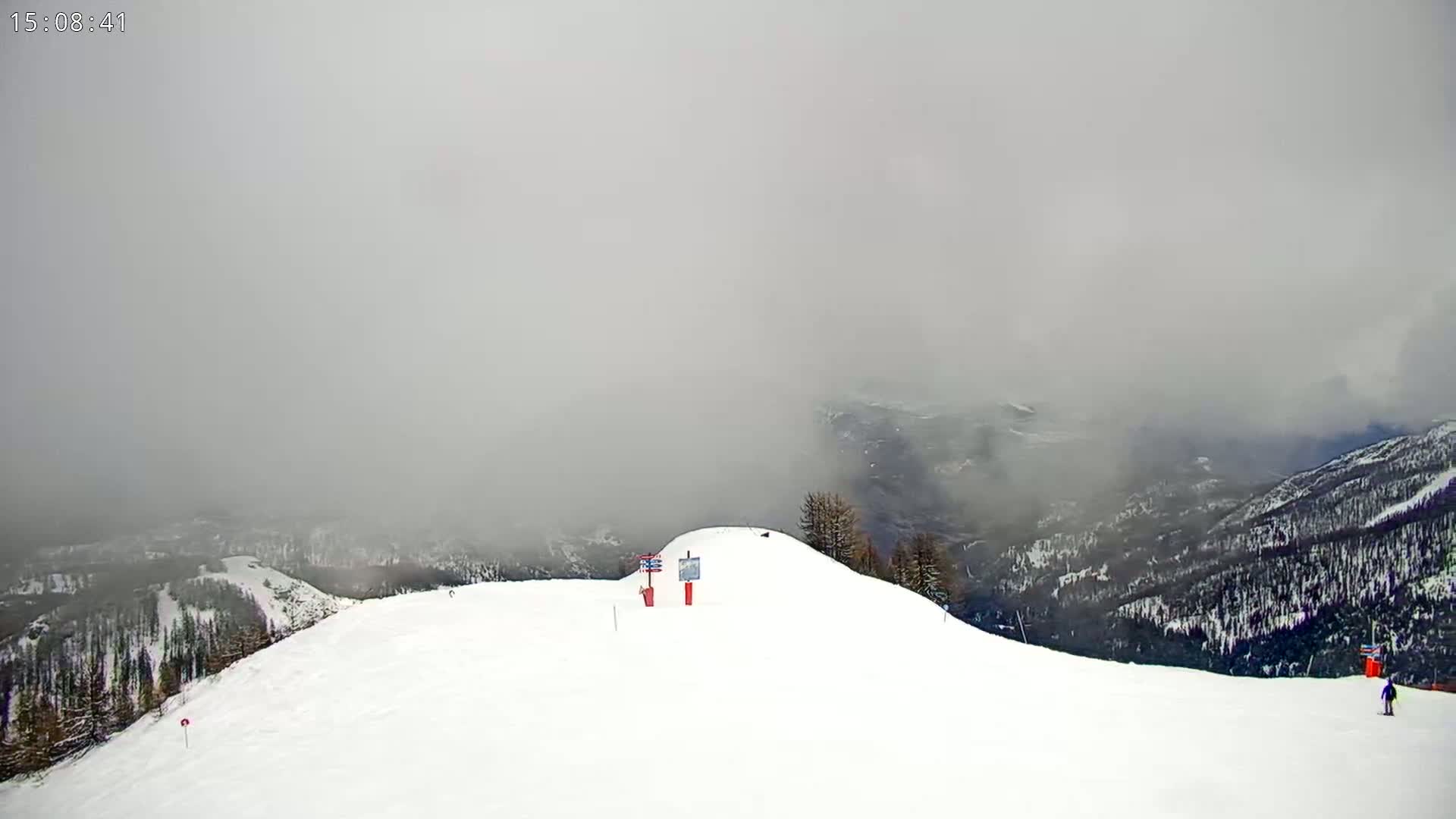A snowy, fog-shrouded mountain slope shows a few skiers and a signpost.