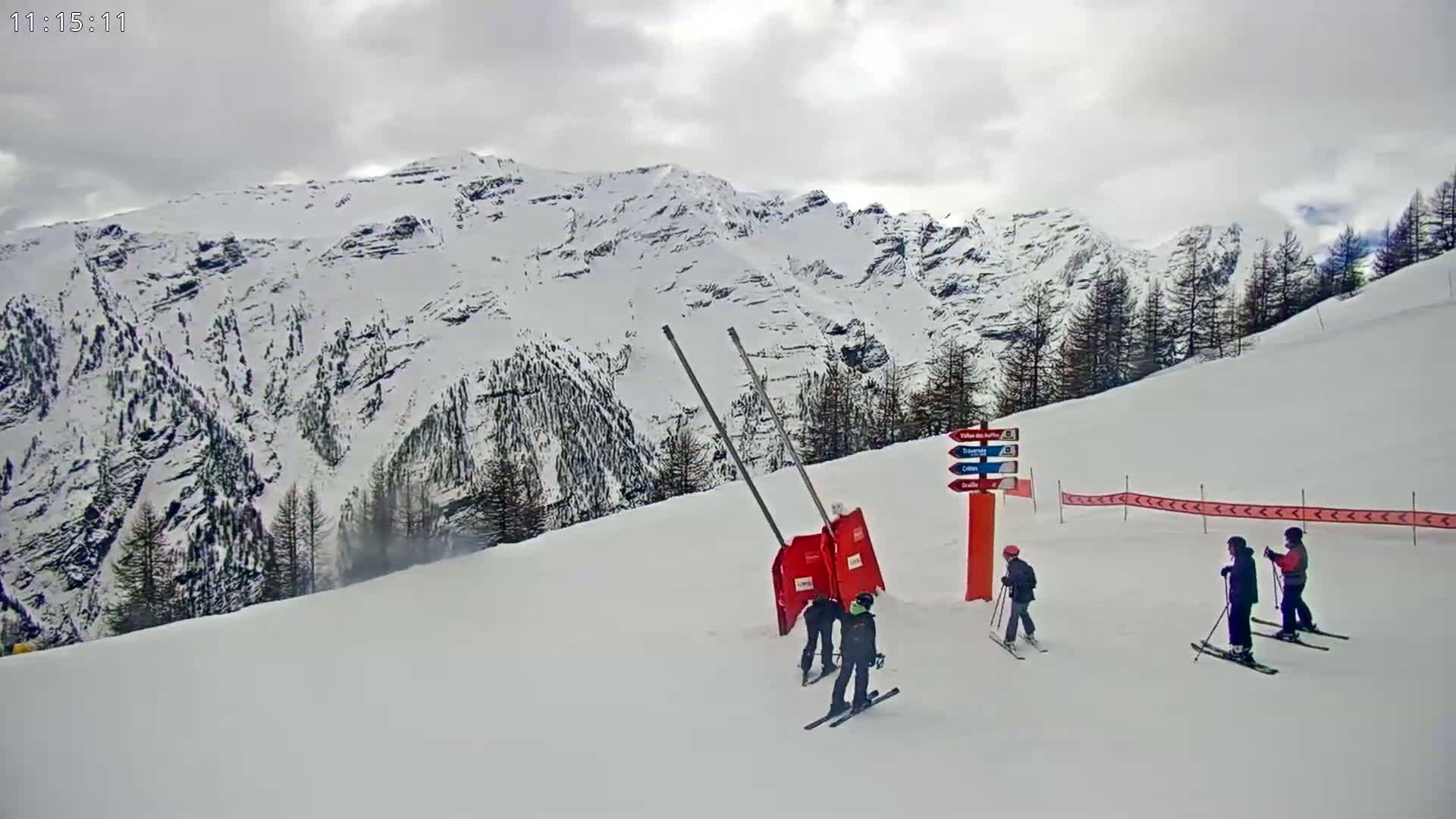 Several skiers are on a snowy mountain slope under a cloudy sky, with snow-covered mountains in the background.