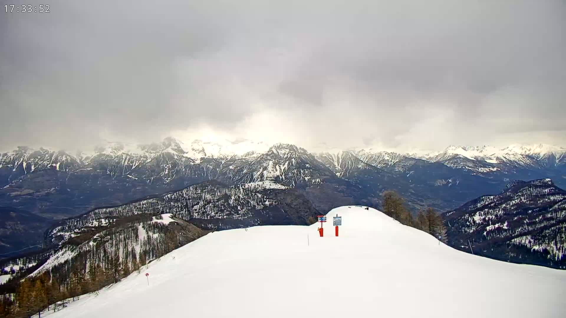 A snow-covered mountain slope overlooks a valley and distant, snow-capped mountain range under an overcast sky.