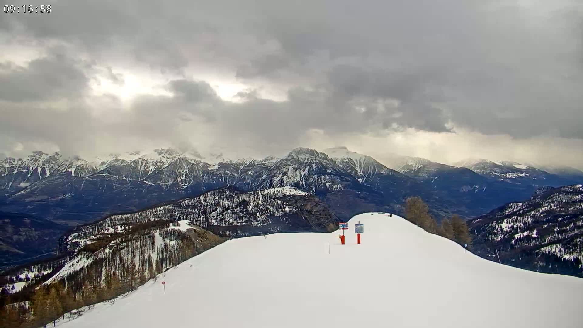 A snow-covered mountain slope overlooks a valley and distant, snow-capped mountain range under a cloudy sky.