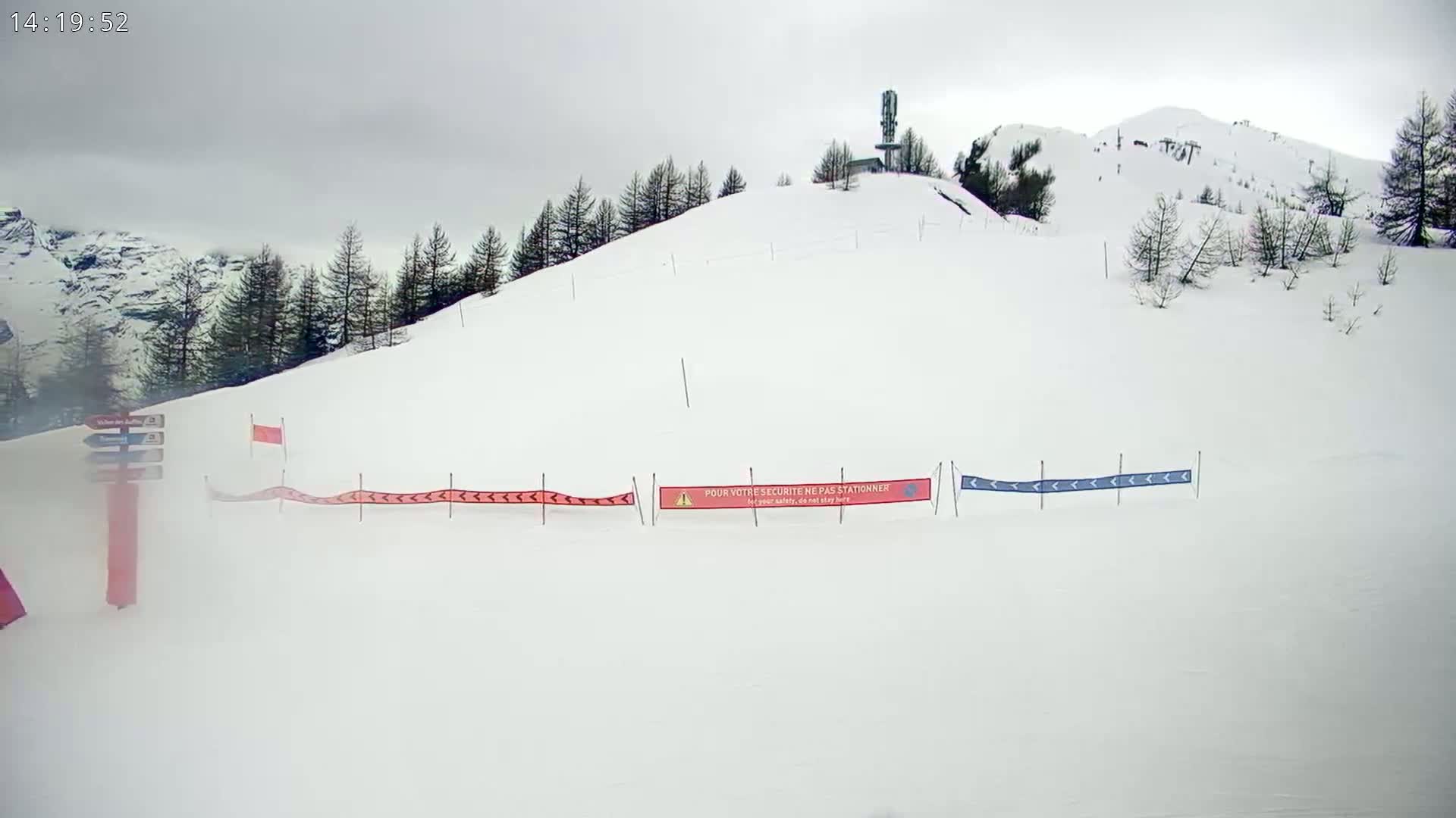 A snow-covered ski slope with a few evergreen trees and a structure atop a hill under an overcast sky.