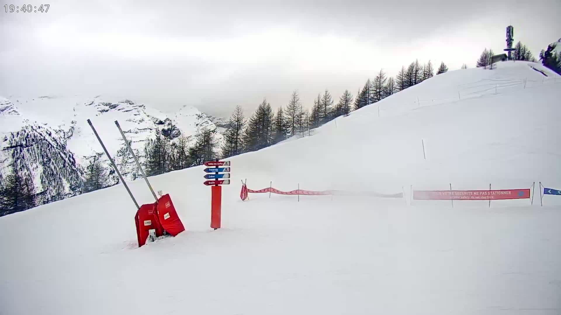 A snowy mountain slope with a few trees, a red signpost, and a red safety barrier is visible under overcast skies.