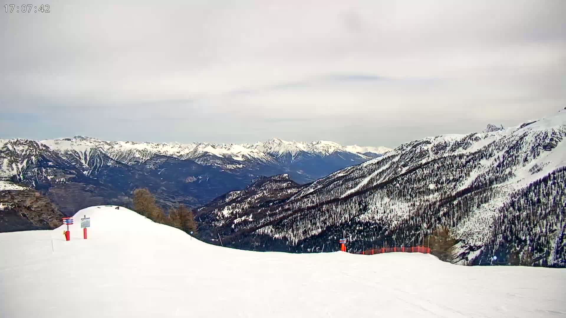 A snow-covered ski slope overlooks a vast mountain range under a cloudy sky.
