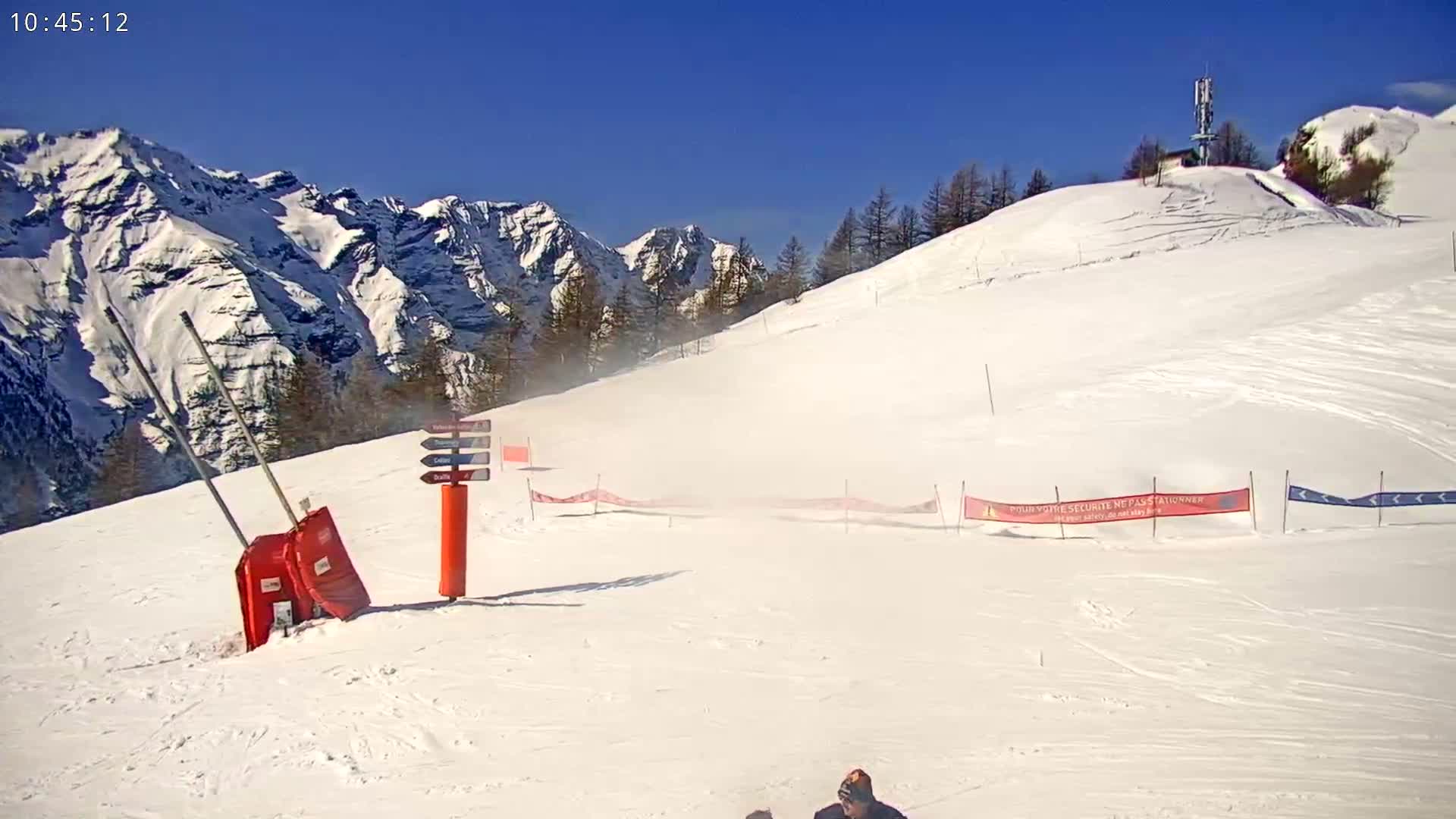 A snow-covered ski slope with a mountainous backdrop under a clear blue sky, showing some light wind blowing snow.