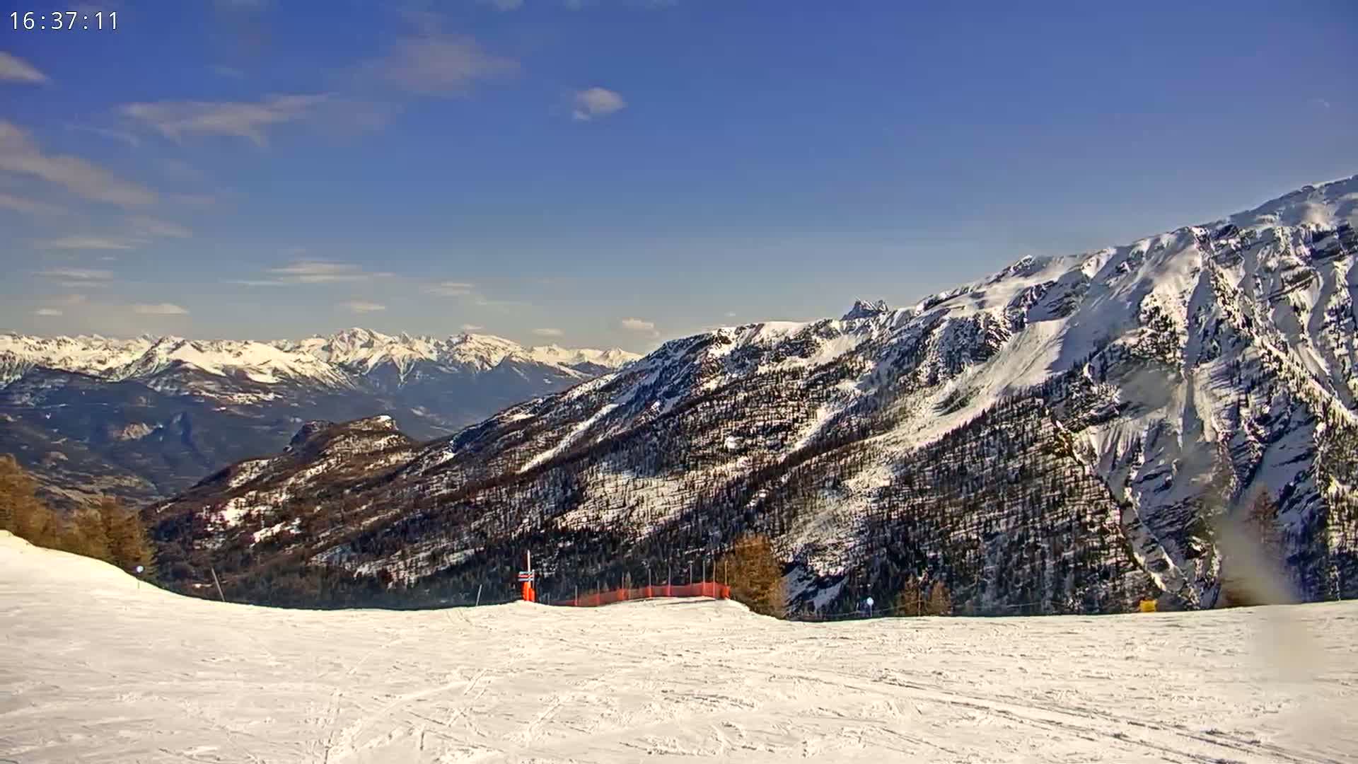 A snow-covered ski slope overlooks a mountain range under a mostly clear, bright blue sky.