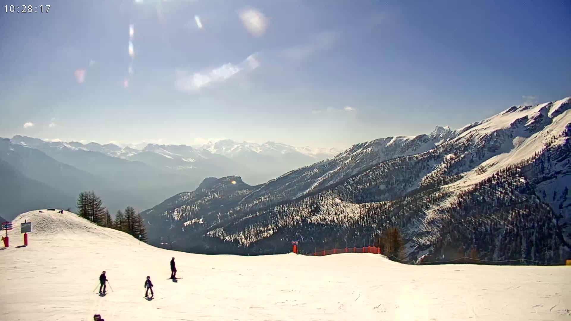A sunny day on a snow-covered mountain slope shows several skiers and snowboarders enjoying the view of a vast mountain range in the distance.