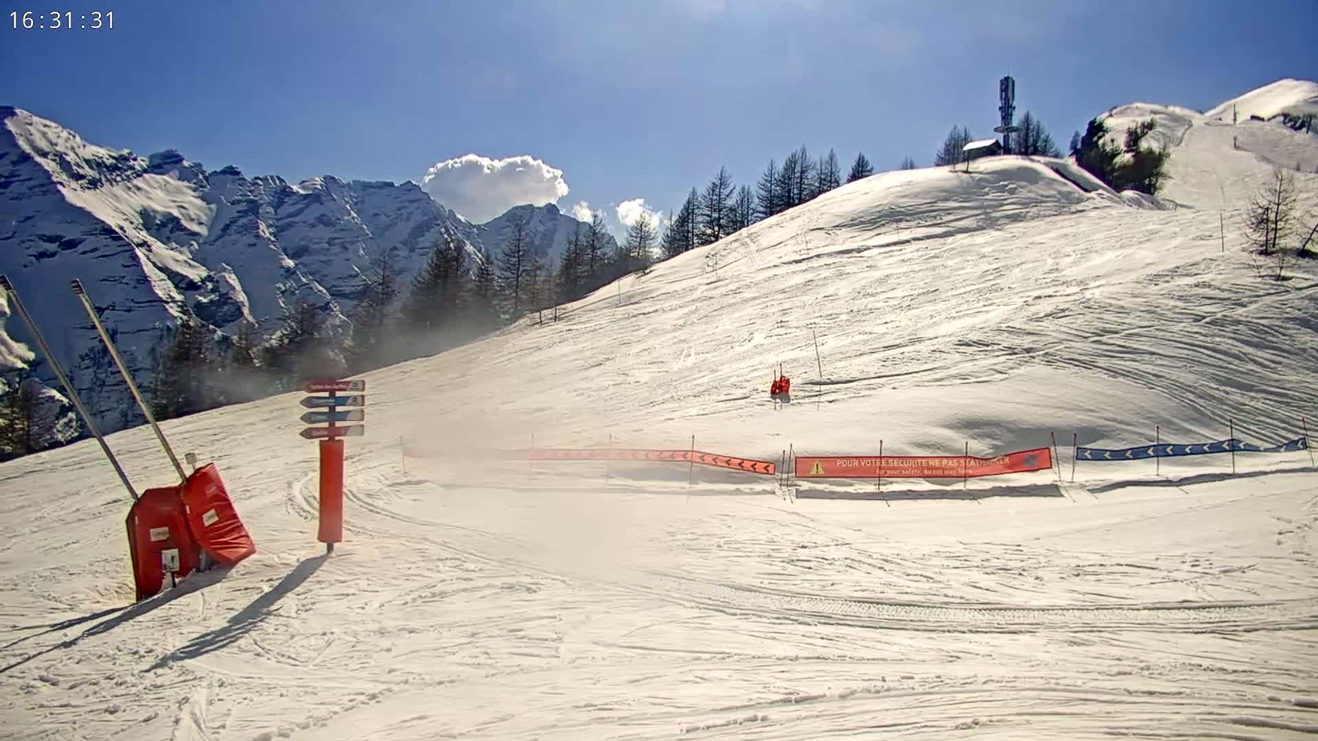 A snowy ski slope with a mountain range in the background under a mostly sunny sky.