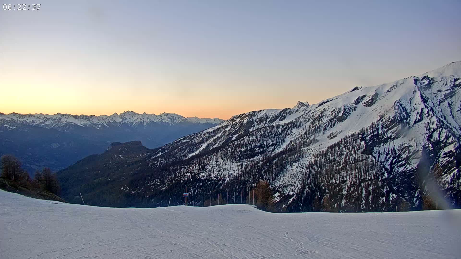 A snowy mountain range at sunset is viewed from a snow-covered slope.