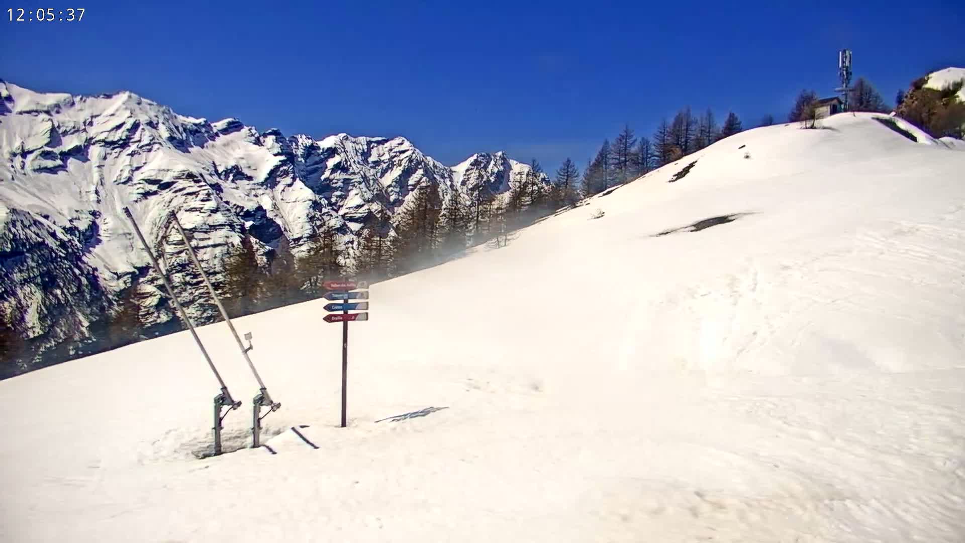 A snow-covered slope with a ski lift and directional signpost, set against a backdrop of snow-capped mountains under a clear blue sky.