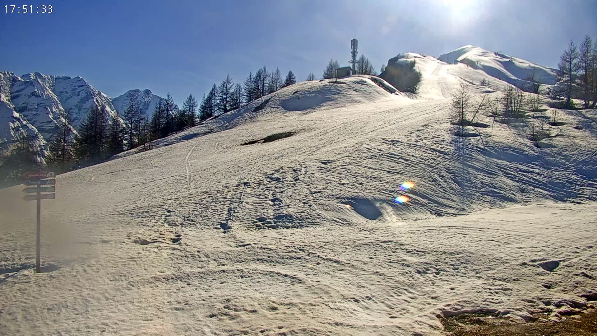A snow-covered hillside with a few trees and a communications tower, under a bright, sunny sky, overlooks a mountain range in the distance.
