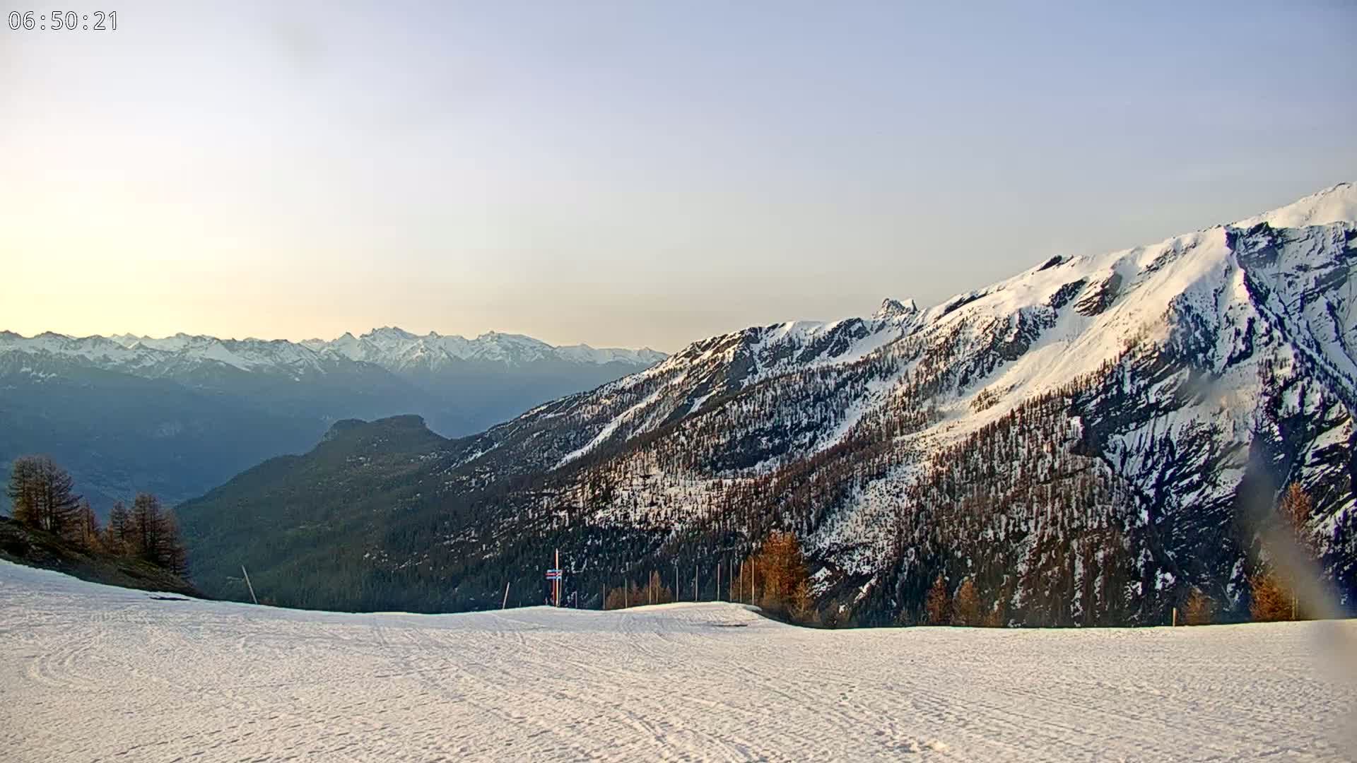 A snow-covered ski slope overlooks a mountain range under a clear, pale sky at dawn.