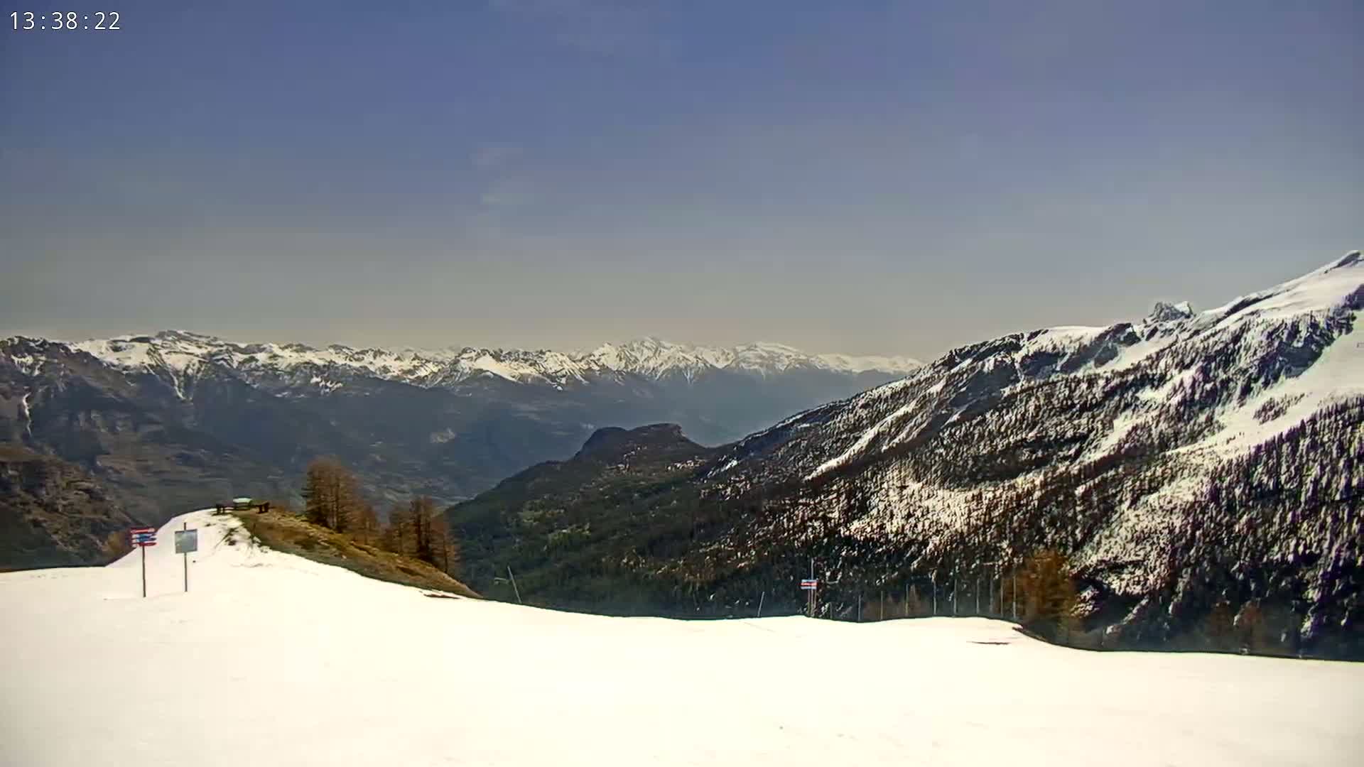 A snow-covered mountain slope overlooks a valley of dark green trees and distant snow-capped mountains under a clear, bright sky.