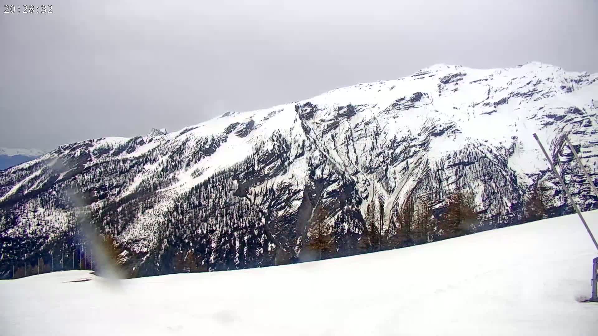 A snow-covered mountain range is viewed from a snowy plain under an overcast sky.