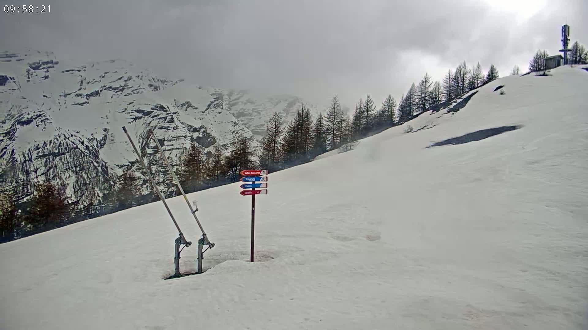 A snow-covered mountain slope with a ski lift mechanism and directional signs, under an overcast sky.