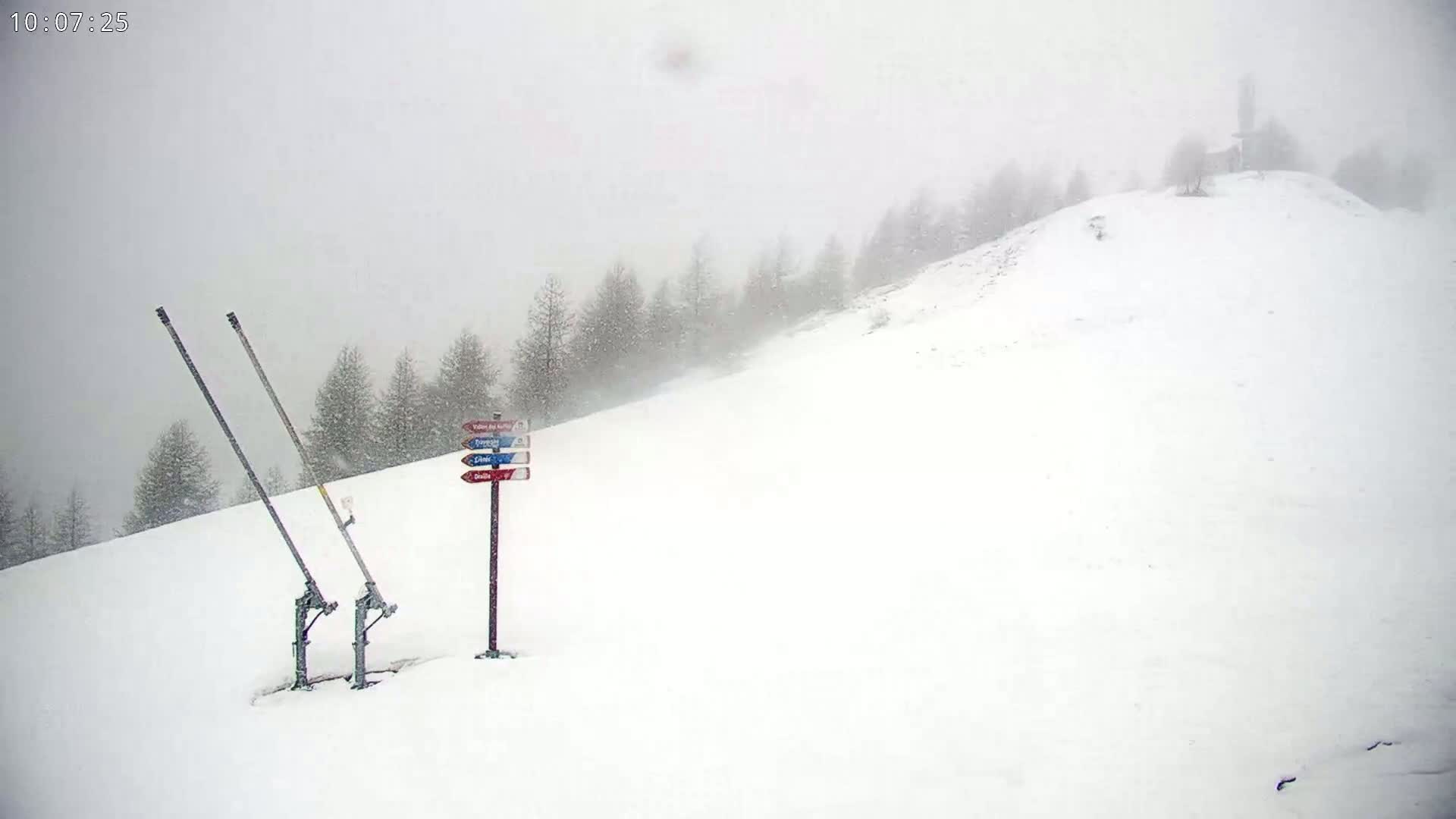 A snowy, overcast landscape shows a ski lift and directional signage on a snow-covered hillside with sparsely wooded trees in the background.