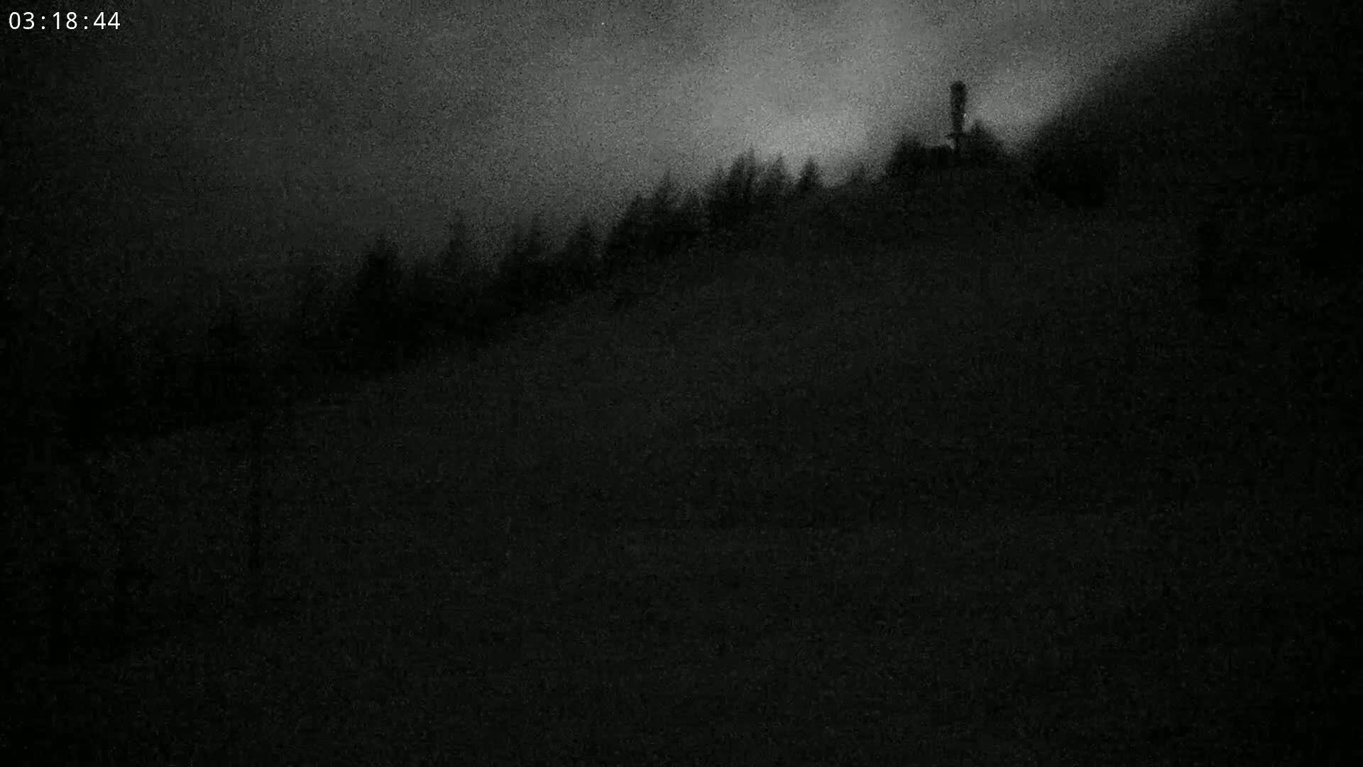 A dark, nighttime view shows a hill covered in silhouetted trees under a cloudy sky.