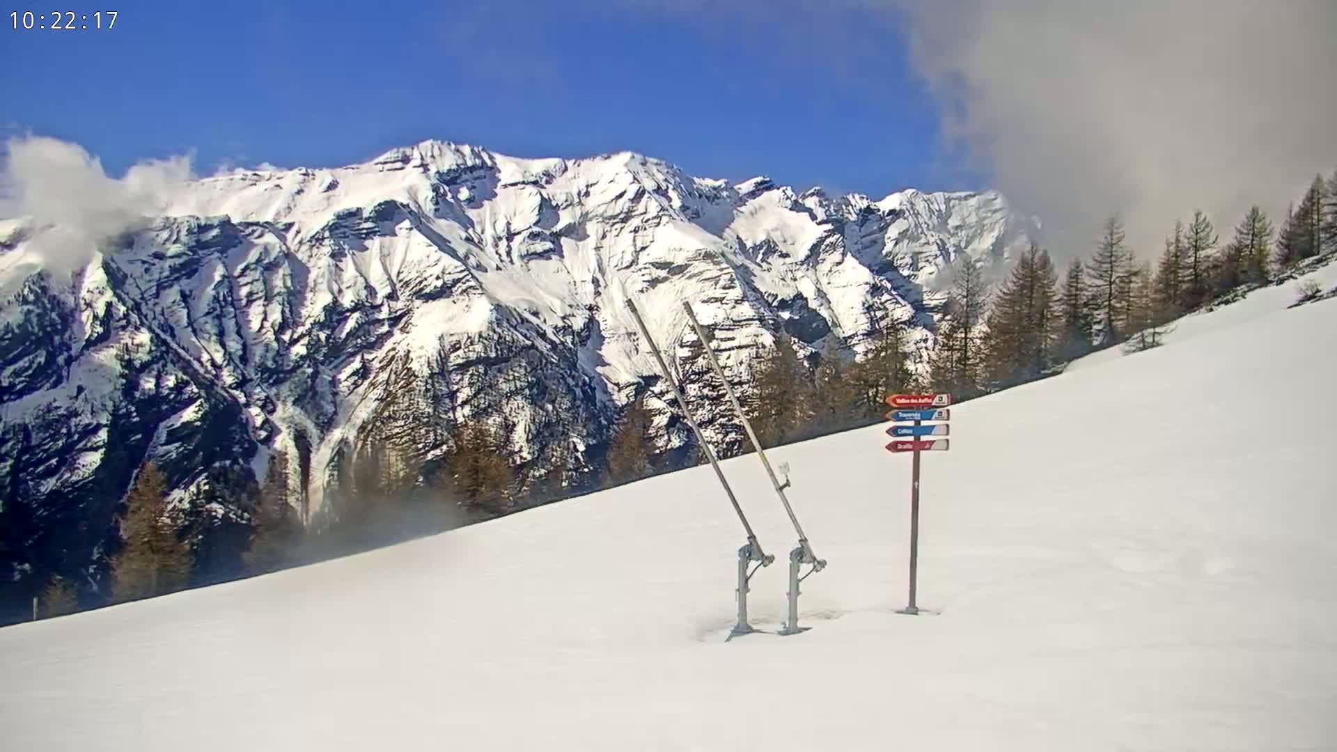 A snowy mountain slope with a ski lift and trail markers under a partly cloudy blue sky.