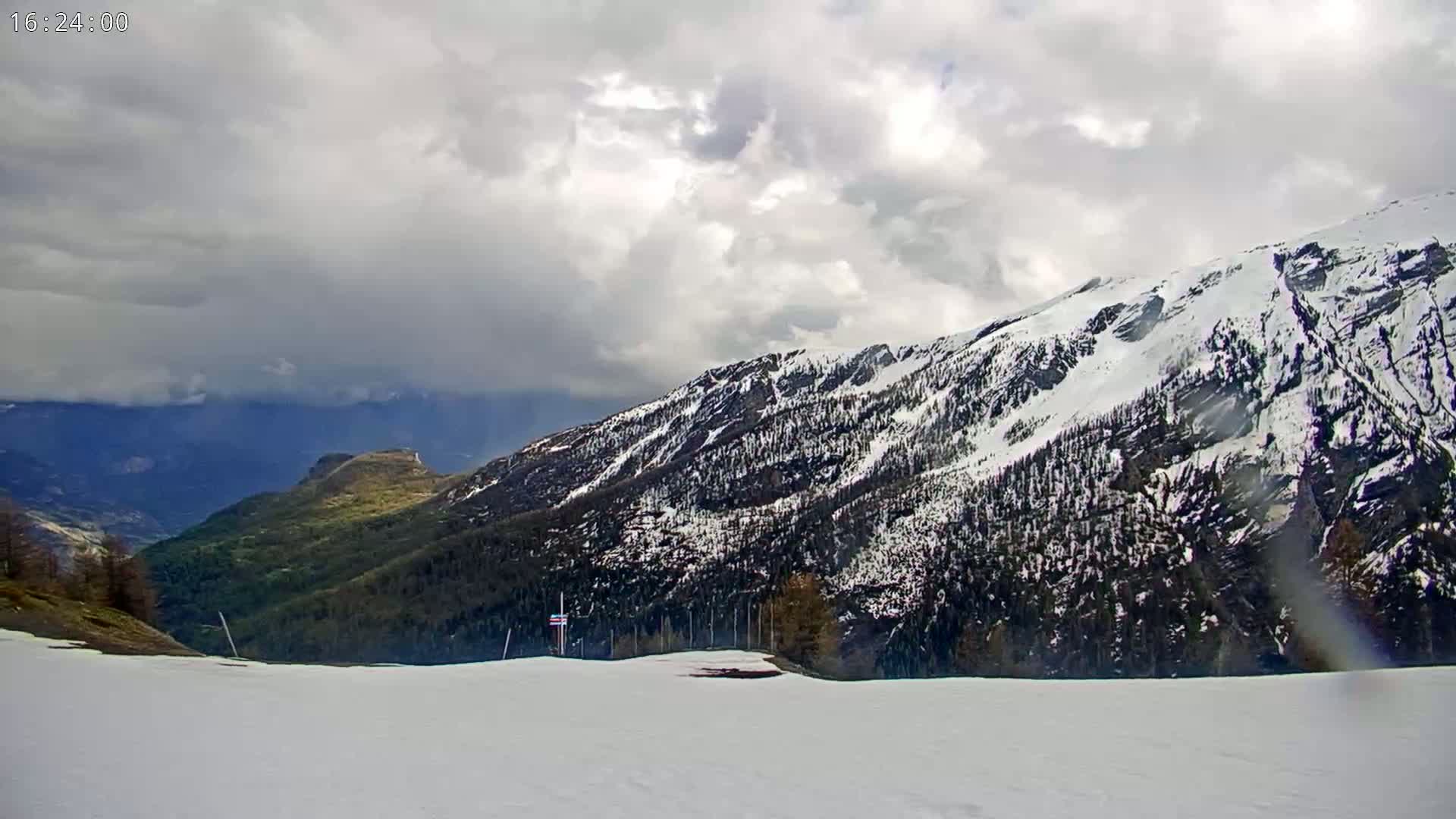 A snow-covered mountainside with dark evergreens is visible under a cloudy sky.