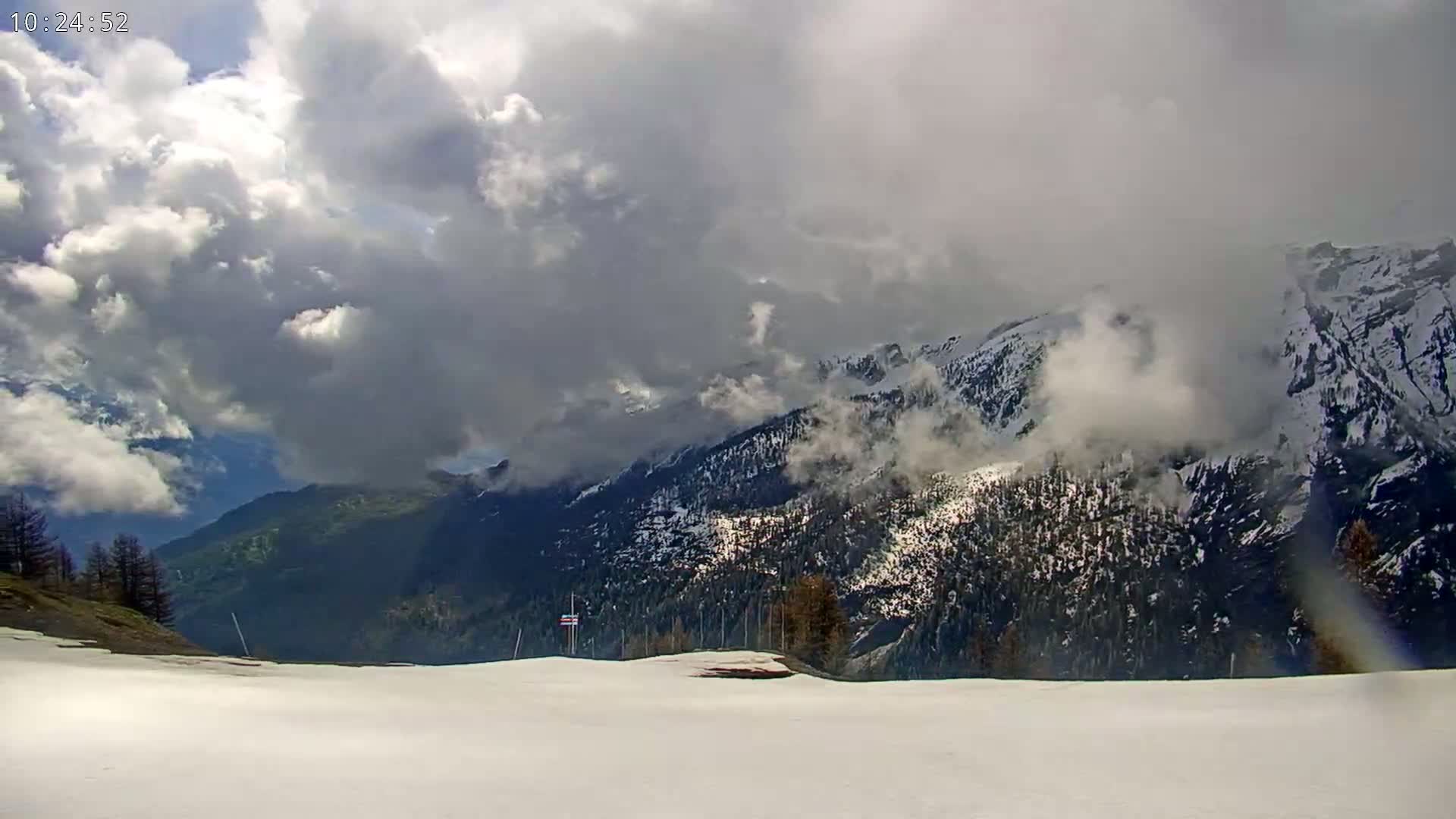 A snow-covered landscape overlooks a mountain range partially obscured by clouds under a partly cloudy sky.