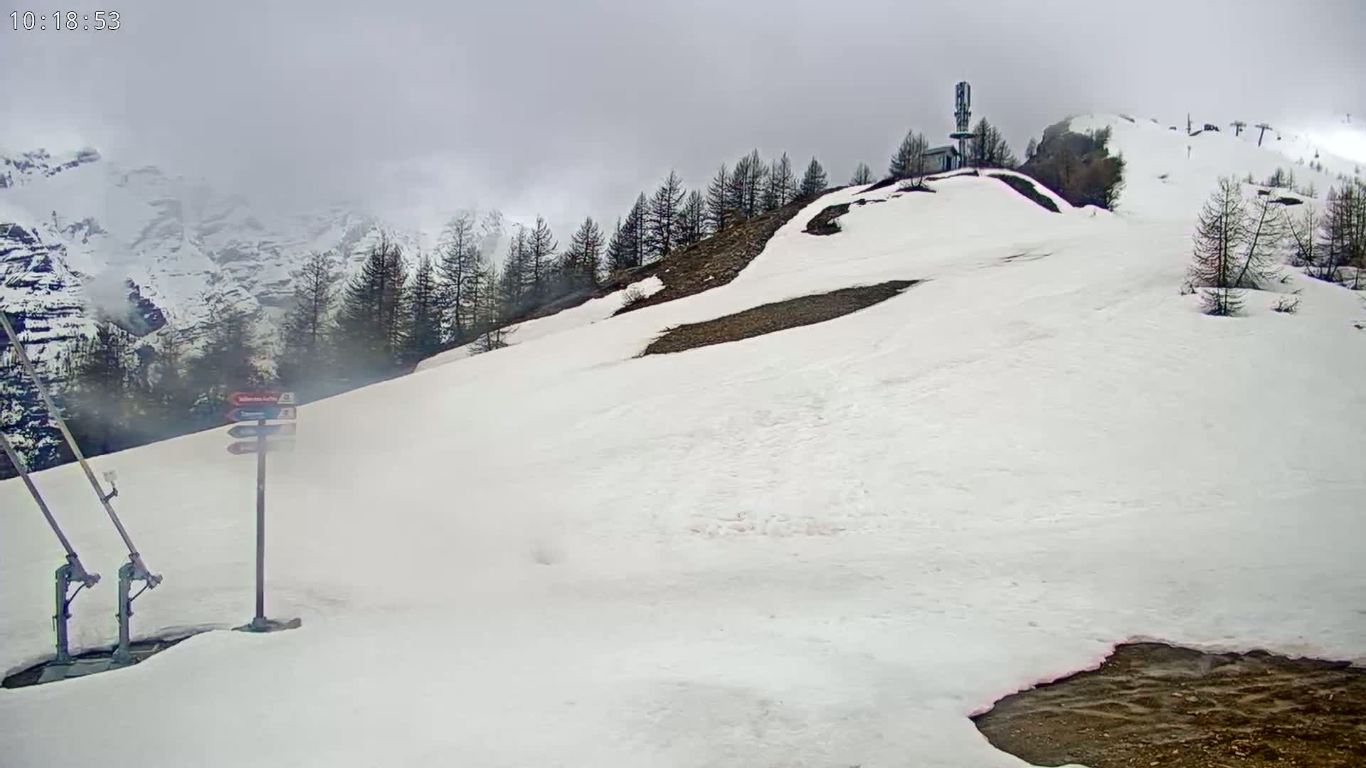 A snow-covered mountain slope with a few evergreen trees, a small building, and a ski lift under an overcast sky.
