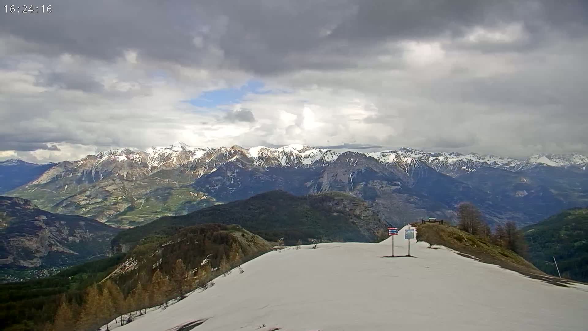 A snow-covered mountaintop overlooks a valley and a distant range of snow-capped mountains under a cloudy sky.