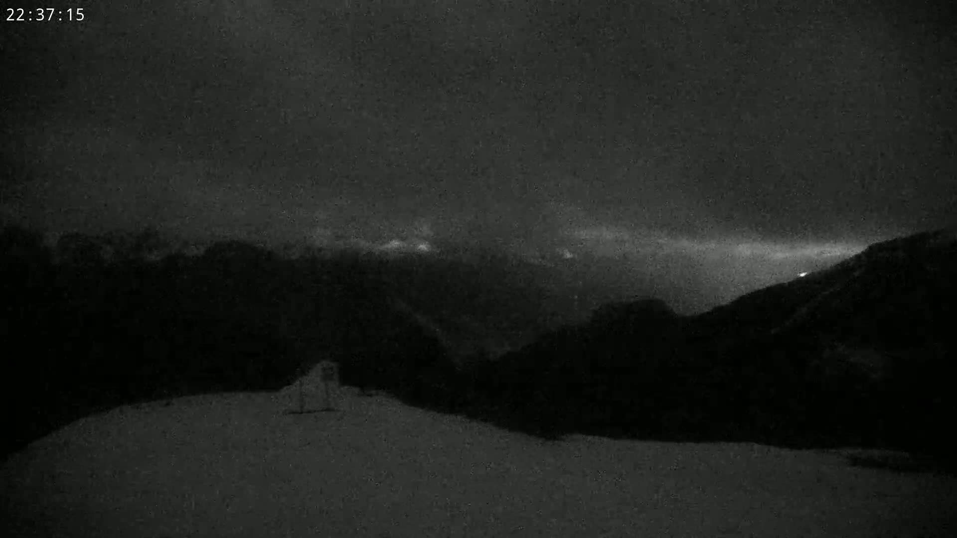 A dark, snowy landscape is shown at night, with a mountain range faintly visible in the distance under a cloudy sky.
