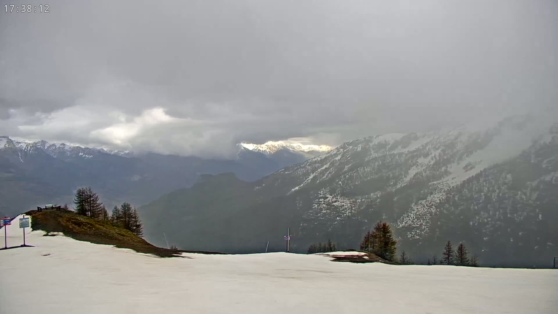 A snow-covered mountain slope overlooks a valley of dark, snow-dusted mountains under a cloudy, overcast sky.