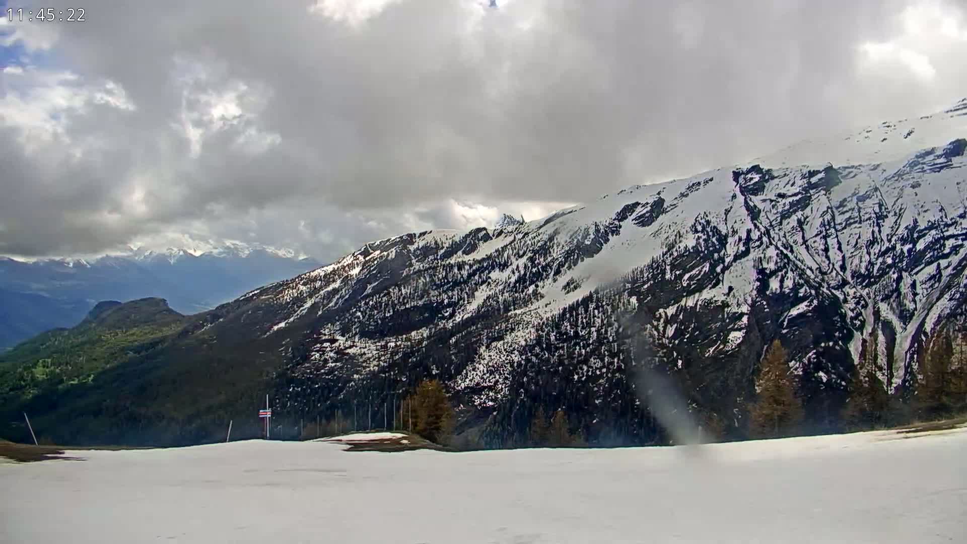 A snow-covered mountain range is visible under a cloudy sky, with a foreground of a mostly snow-covered valley.