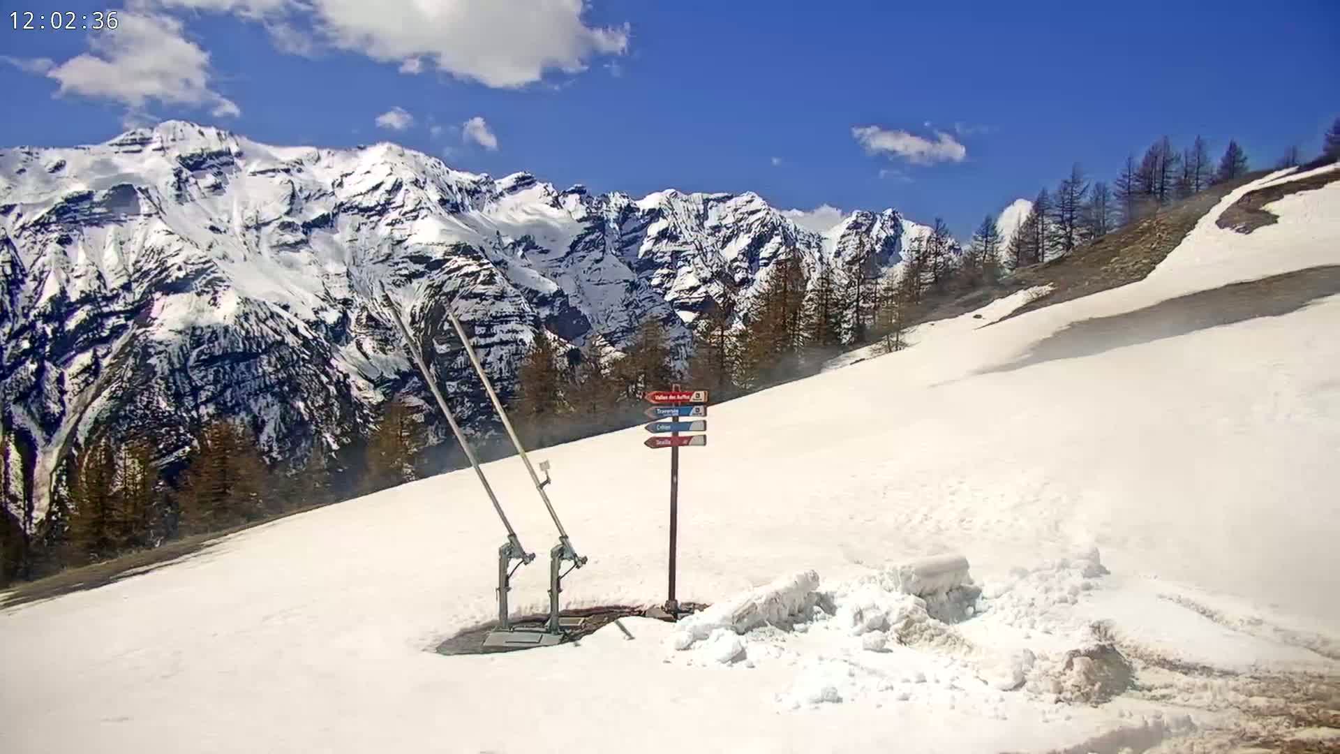A snow-covered mountain slope with a directional sign and ski lift equipment is visible under a partly cloudy blue sky.