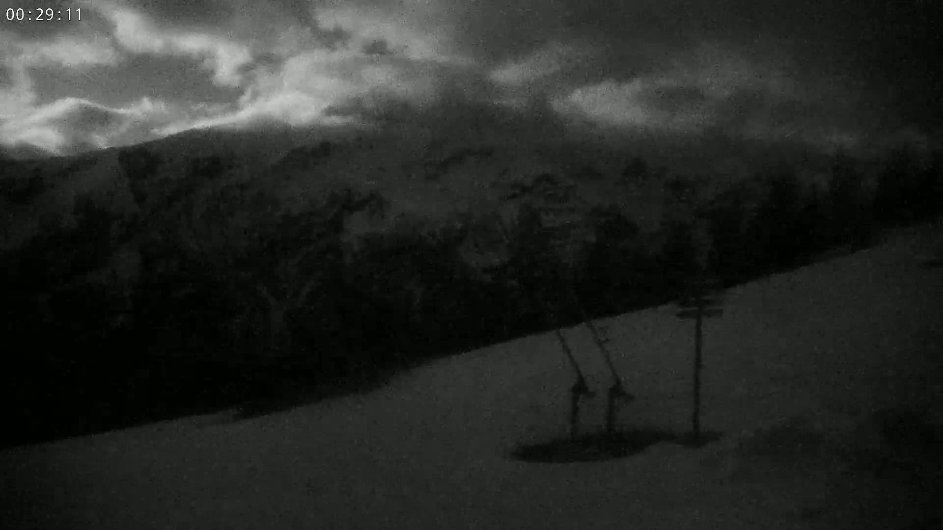 A nighttime, snowy, mountainous landscape is shown, with two ski poles lying in the snow in the foreground under a cloudy sky.