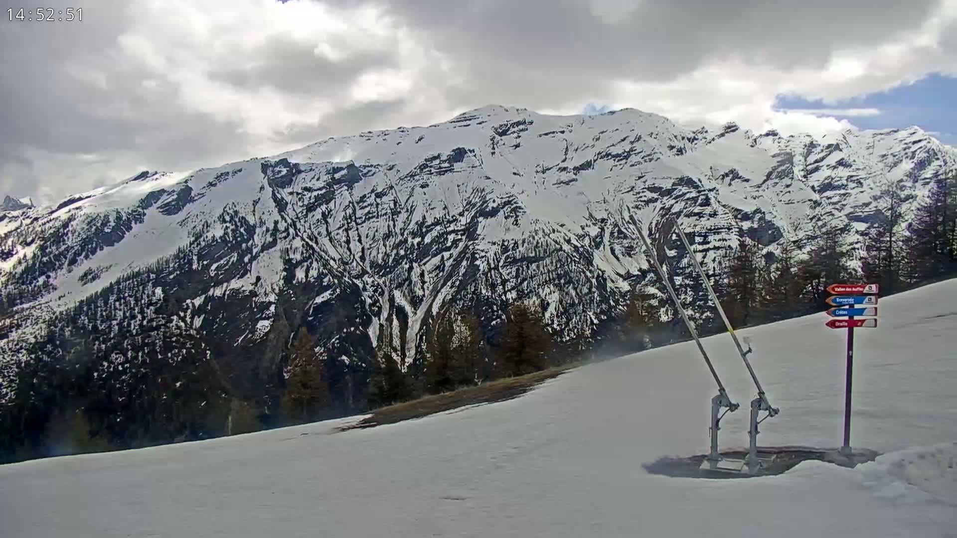 A snow-covered mountain range is visible under a cloudy sky, with a ski lift mechanism in the foreground on a snowy slope.