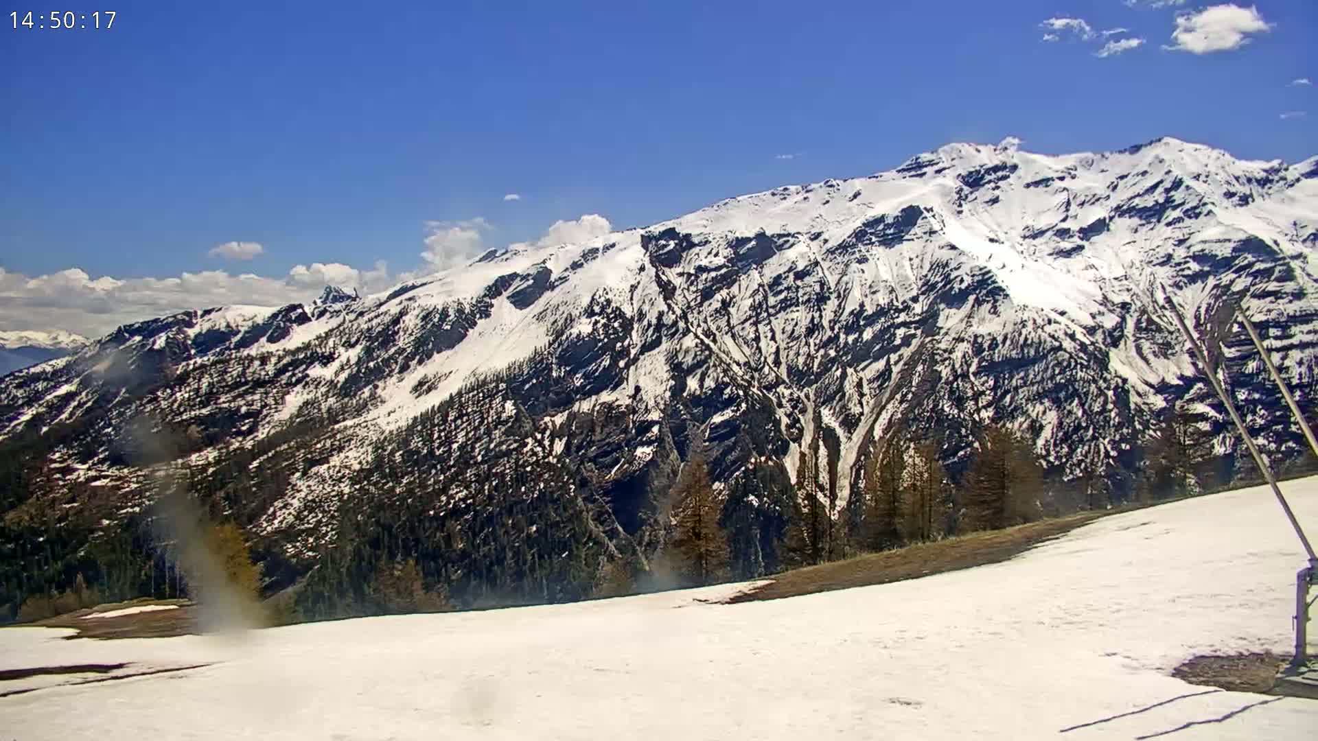 A snow-covered mountain range is visible under a mostly clear blue sky, with some patchy snow remaining on the lower slopes.