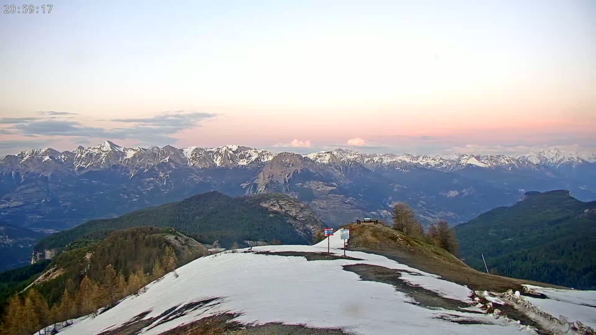 A snow-dusted mountaintop overlooks a vast range of snow-capped mountains under a pale pink and purple sunset sky.