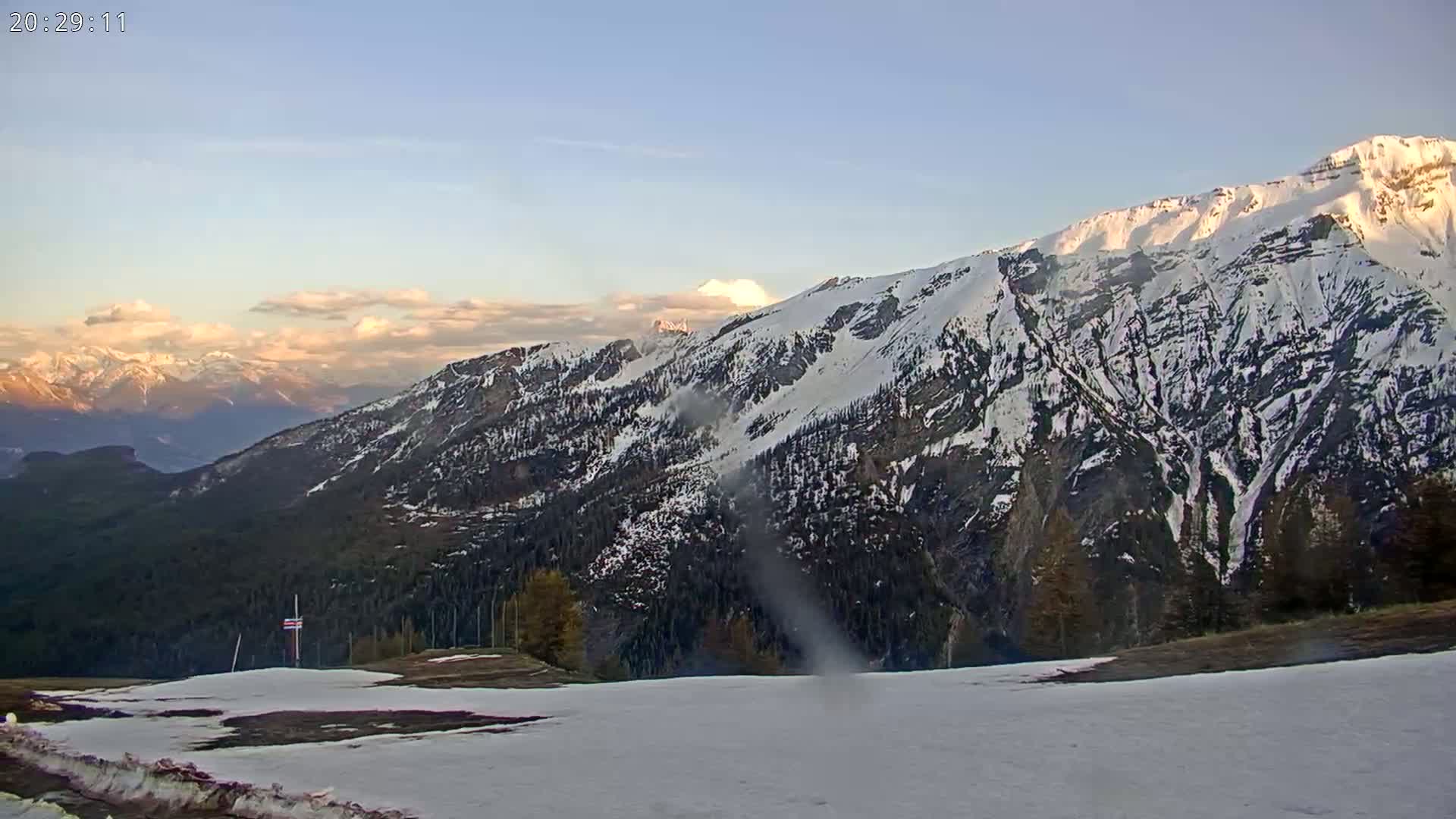 A snow-covered mountain range at sunset, with a partially snow-covered foreground and a clear sky.
