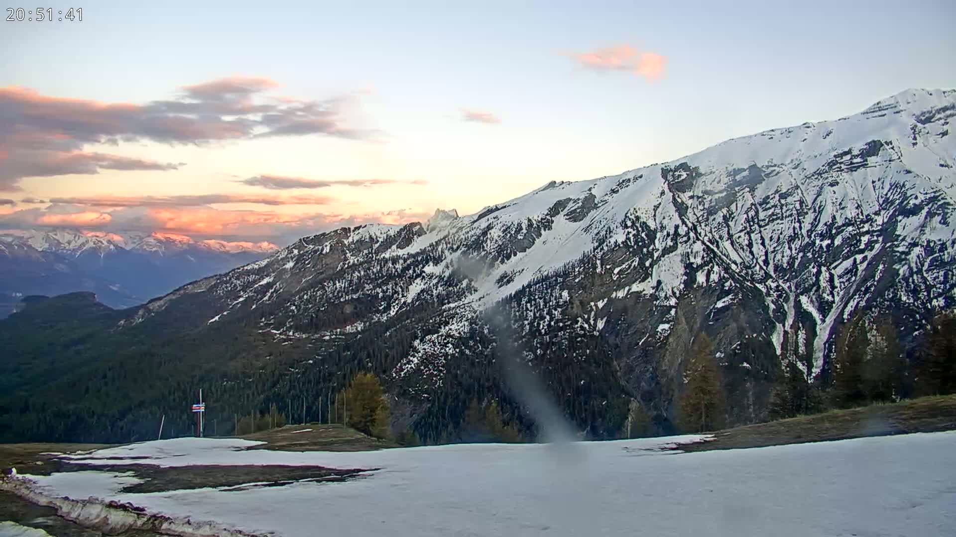 A snow-covered mountain range at sunset, with pink clouds in the sky, is viewed from a lower vantage point where some patches of snow remain on the ground.