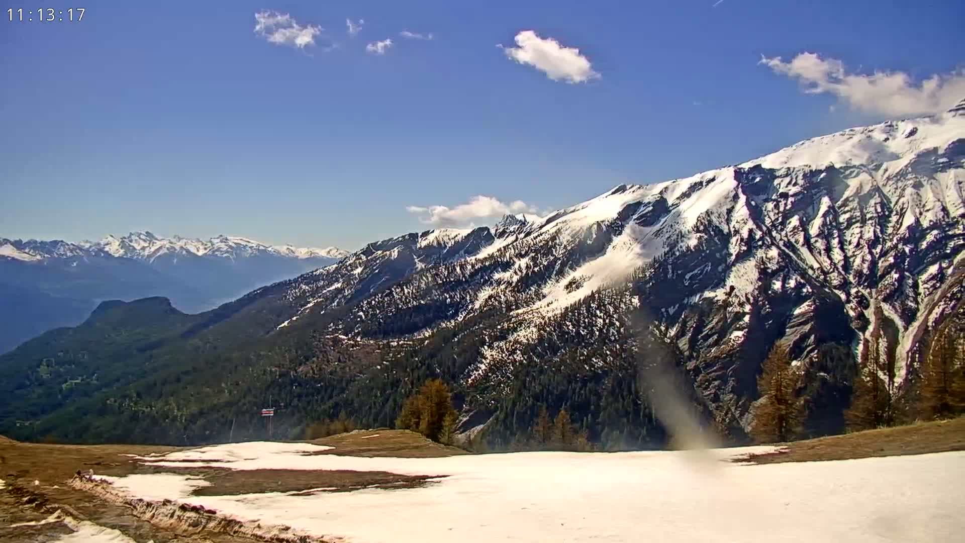 A mostly clear sky overlooks snow-capped mountains and a valley with patches of snow and brown grass in the foreground.