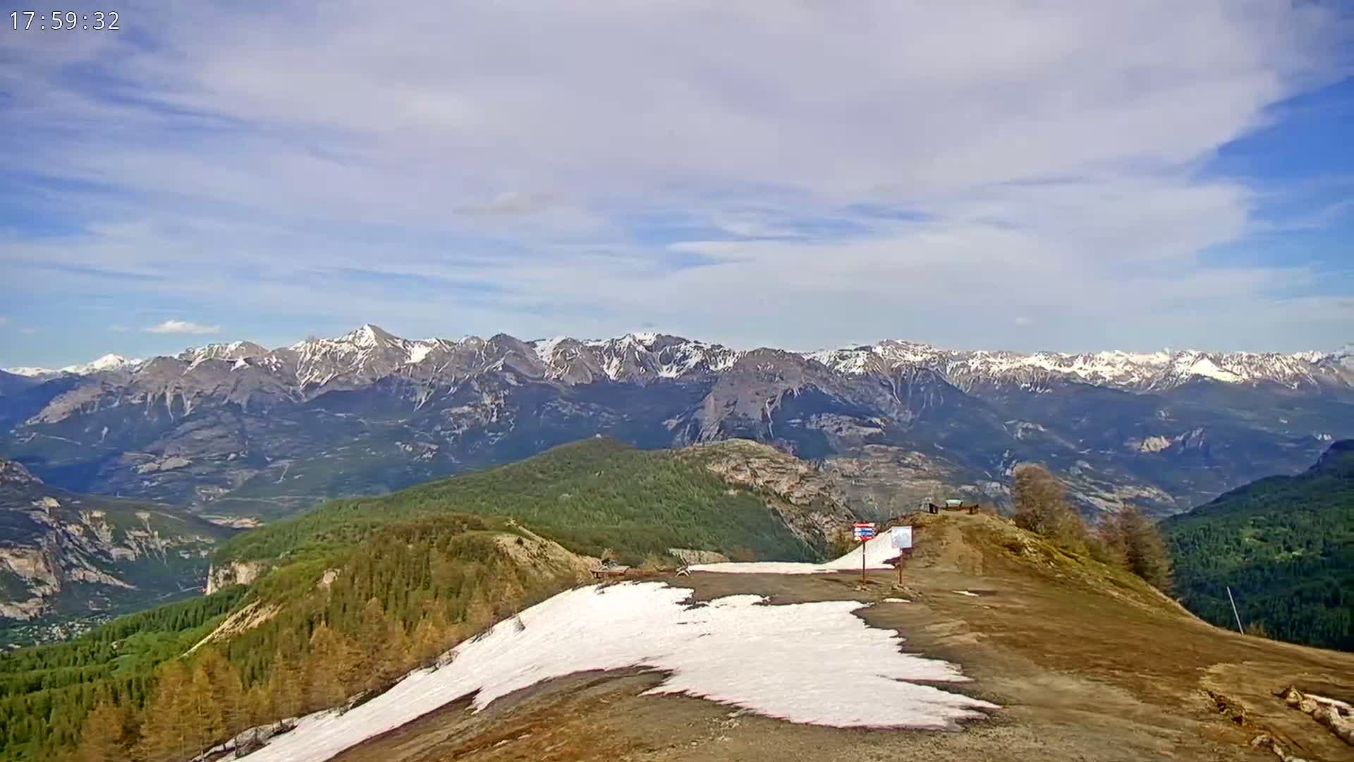 A mountaintop view shows a partly snow-covered hill overlooking a valley and a distant range of snow-capped mountains under a partly cloudy sky.