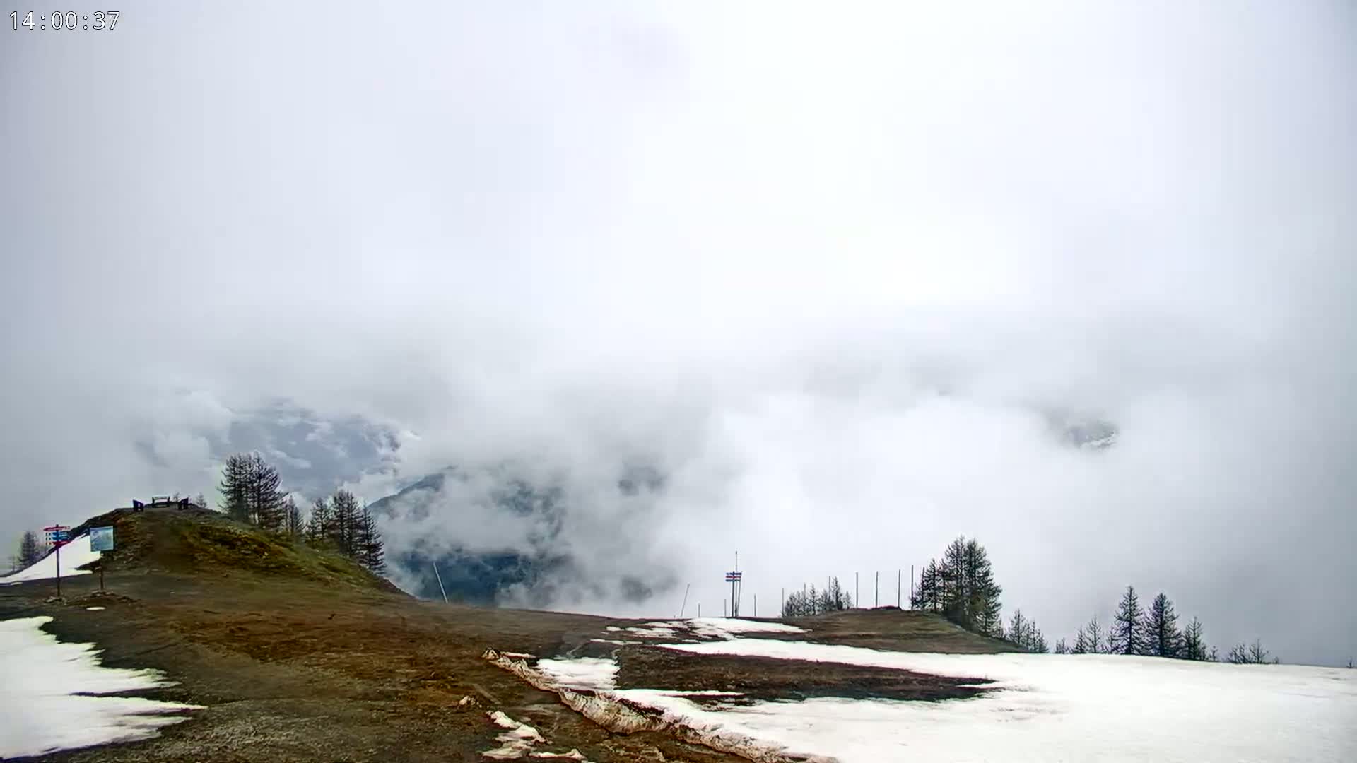 A foggy mountaintop scene with patches of snow and dark green conifers, a few small structures, and some bare, brown earth visible.