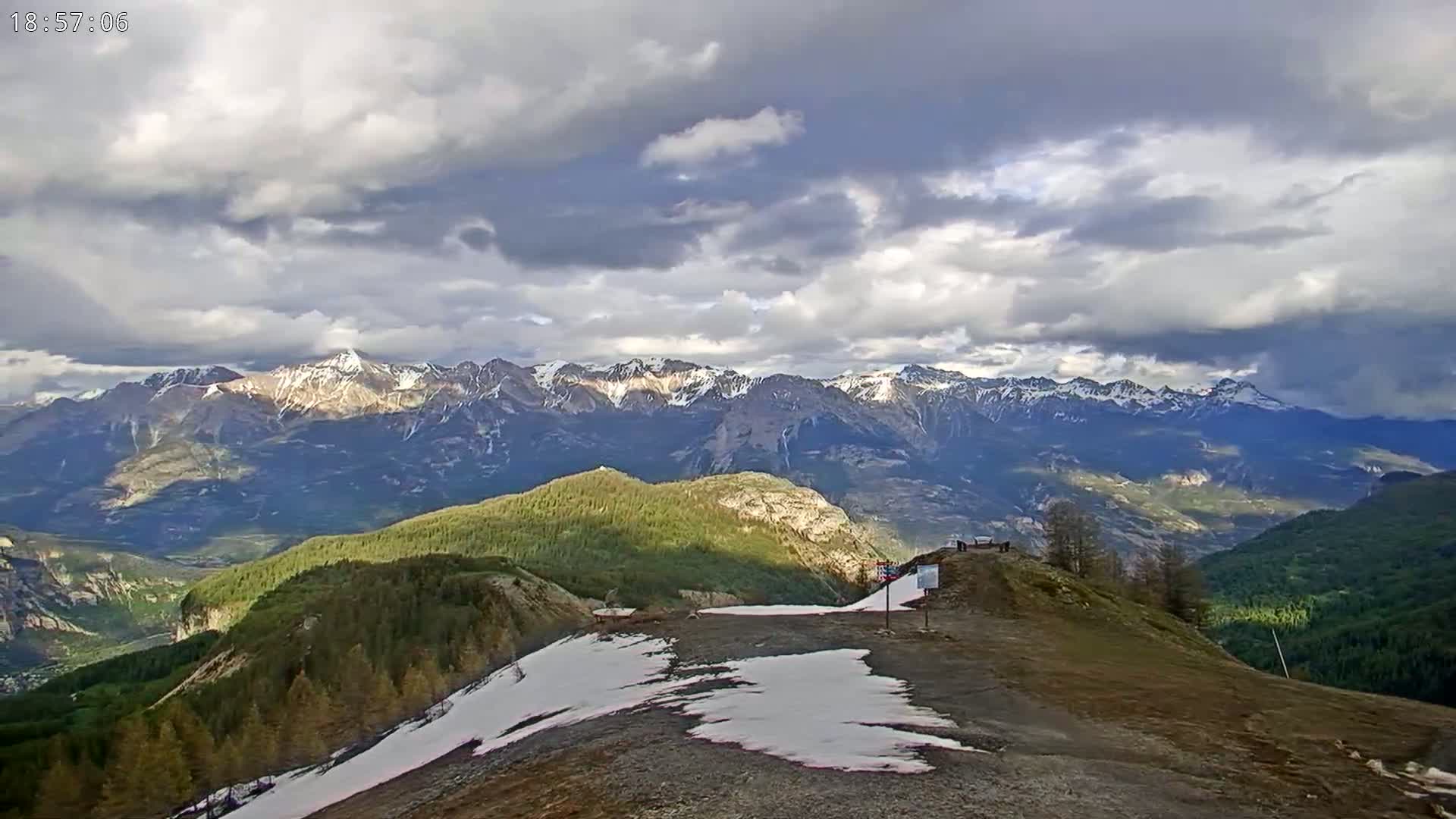 A mountaintop view shows a partly cloudy sky over a valley with snow-capped mountains in the distance, and patches of snow on the ground in the foreground.