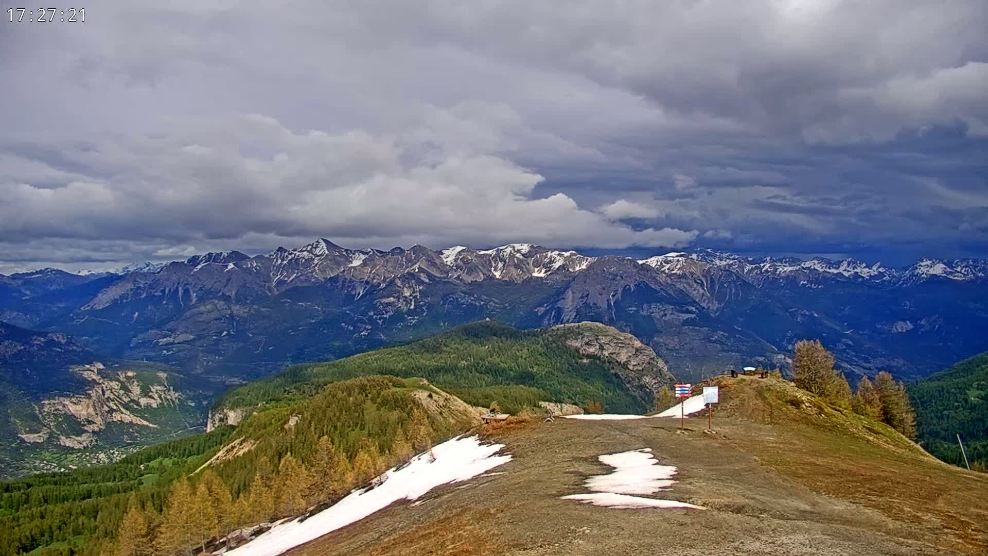 A mountaintop view under a cloudy sky shows a rocky, grassy landscape with patches of snow, overlooking a valley and a distant range of snow-capped mountains.
