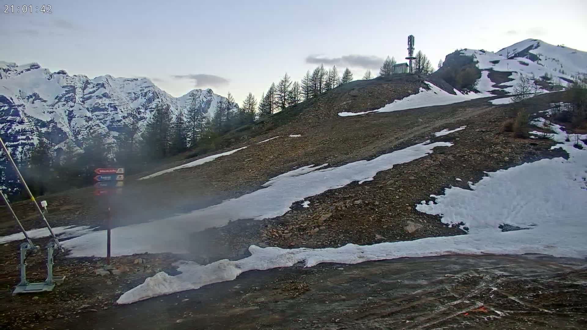 A rocky, snow-dusted hillside with a few evergreen trees, under a clear twilight sky, overlooks snow-capped mountains in the distance.