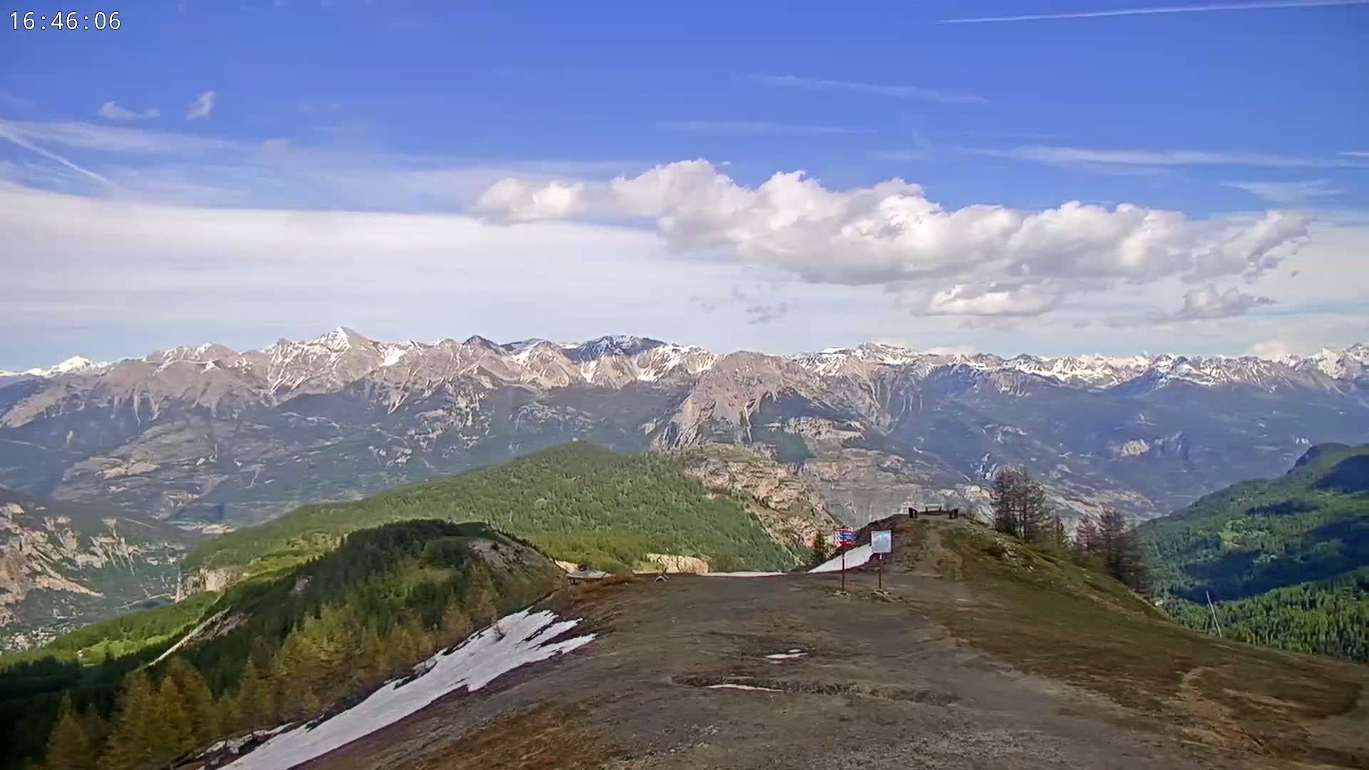 A mountaintop view shows a mostly sunny sky with some clouds, a rocky, sparsely vegetated foreground, a valley of green forests, and a distant mountain range with snow-capped peaks.