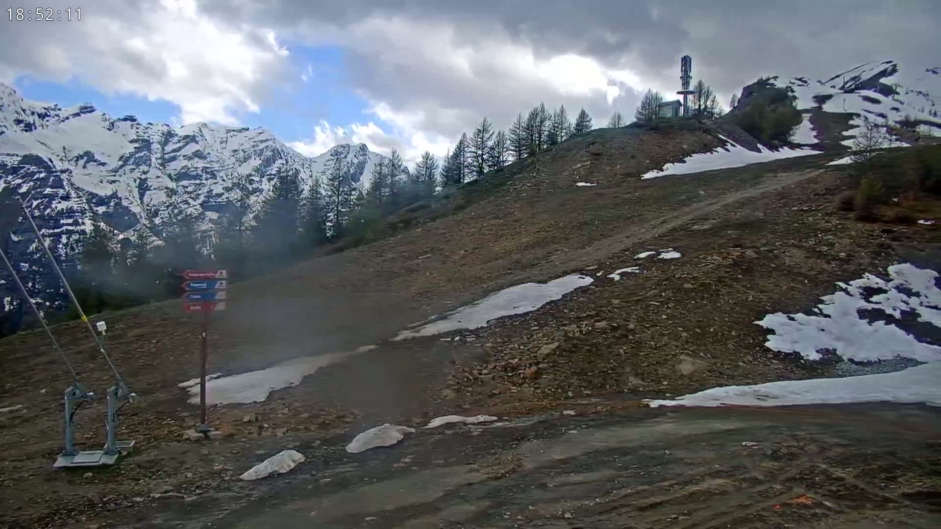 A rocky, snow-dusted hillside slopes down towards a viewer, with snow-capped mountains in the background under a partly cloudy sky.