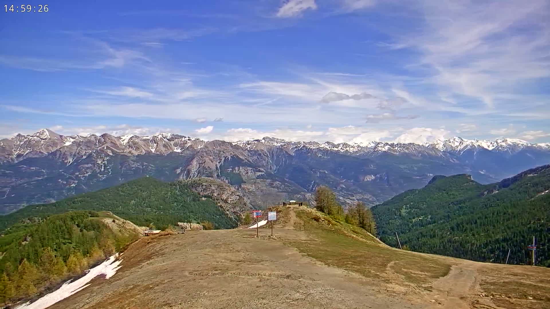 A mostly clear sky overlooks a mountainous landscape with snow-capped peaks in the distance and a grassy, sparsely vegetated hill in the foreground.