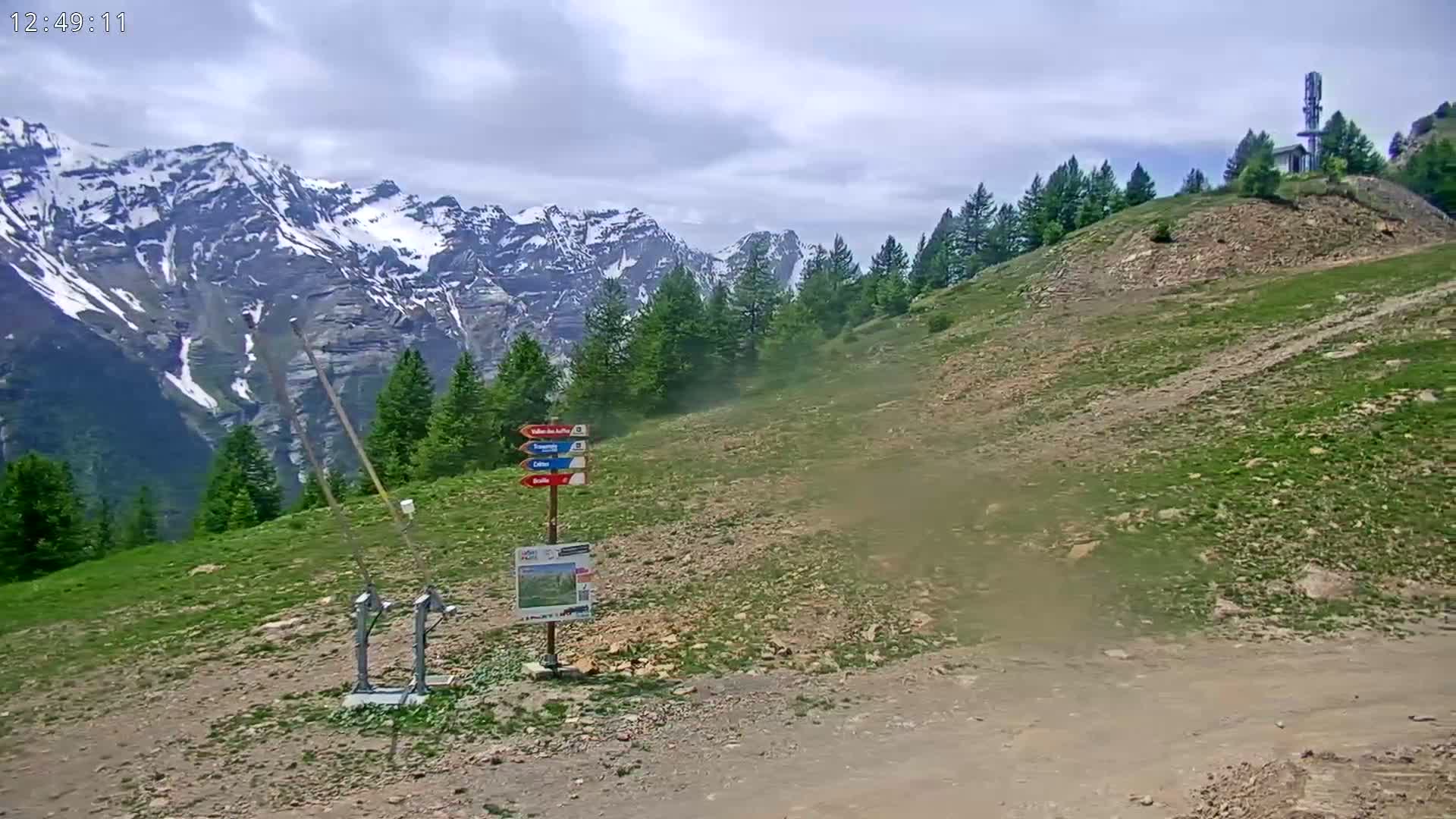 A mountainside with sparse vegetation, a ski lift mechanism, and snow-capped mountains in the background under partly cloudy skies.