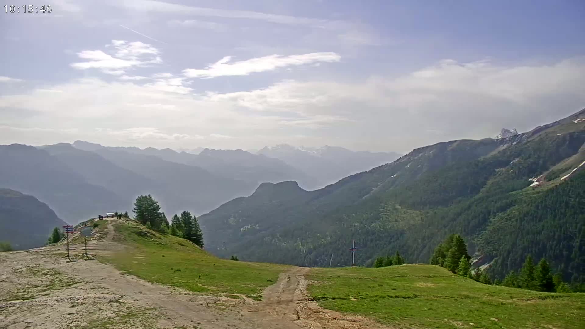 A mountaintop view on a partly cloudy day shows a grassy expanse leading to a dirt path that descends into a valley of rolling green hills and distant, hazy mountains.