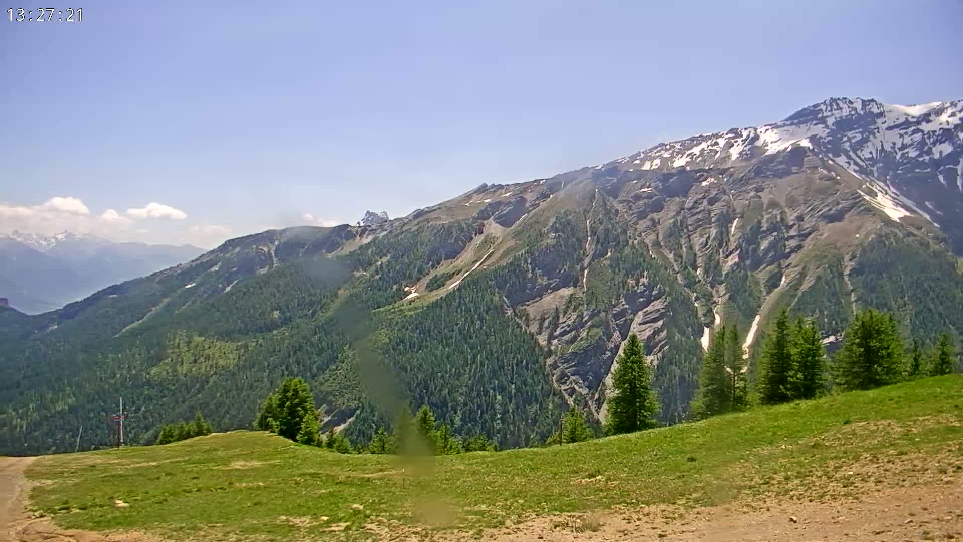 A partly snow-covered mountain range is visible under a mostly clear sky, with a foreground of green grass and a few evergreens.