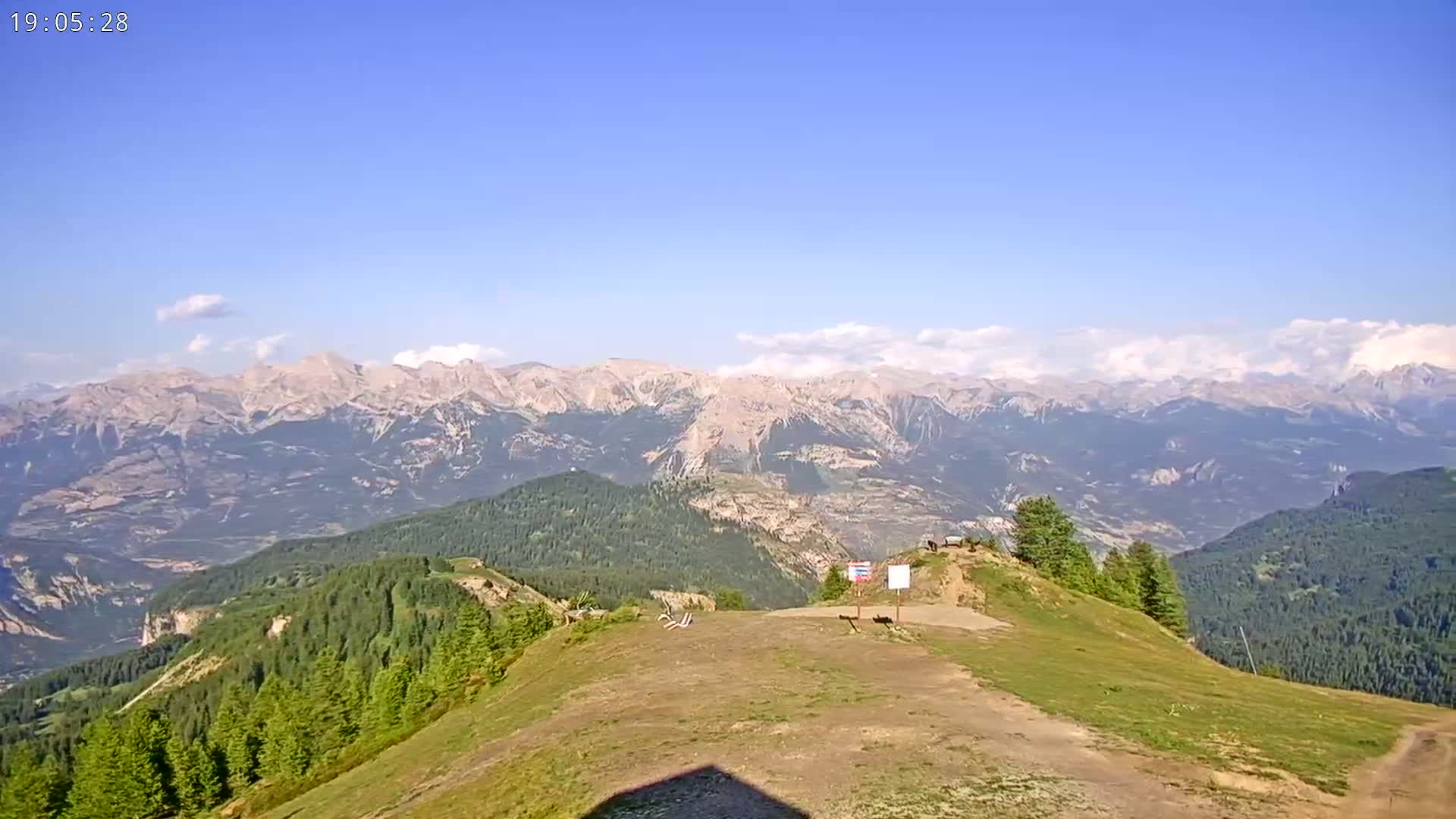 A mountaintop view shows a mostly clear sky, a grassy expanse with a dirt path, evergreen forests below, and a vast range of mountains in the distance.