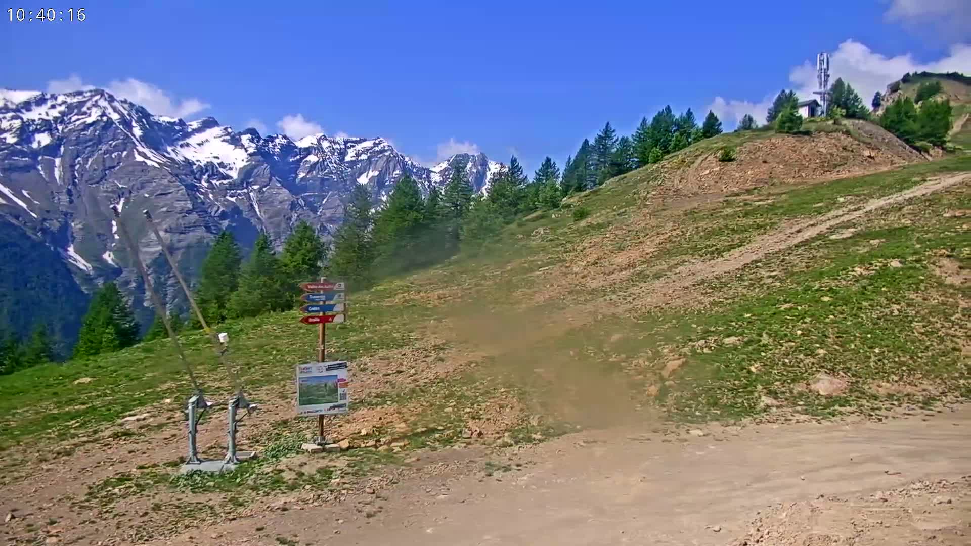 A mostly sunny, dusty mountainside path leads to a snow-capped mountain range under a clear blue sky.
