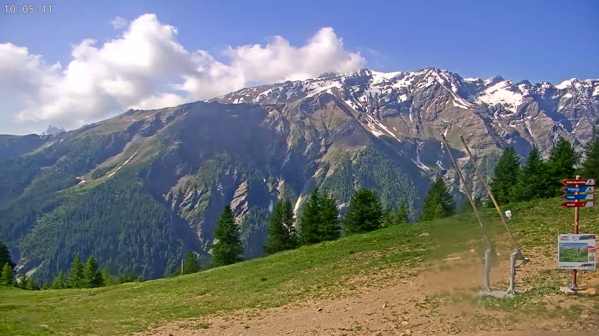 A grassy hillside with a small structure and a mountain range with snow-capped peaks in the background under a partly cloudy sky.